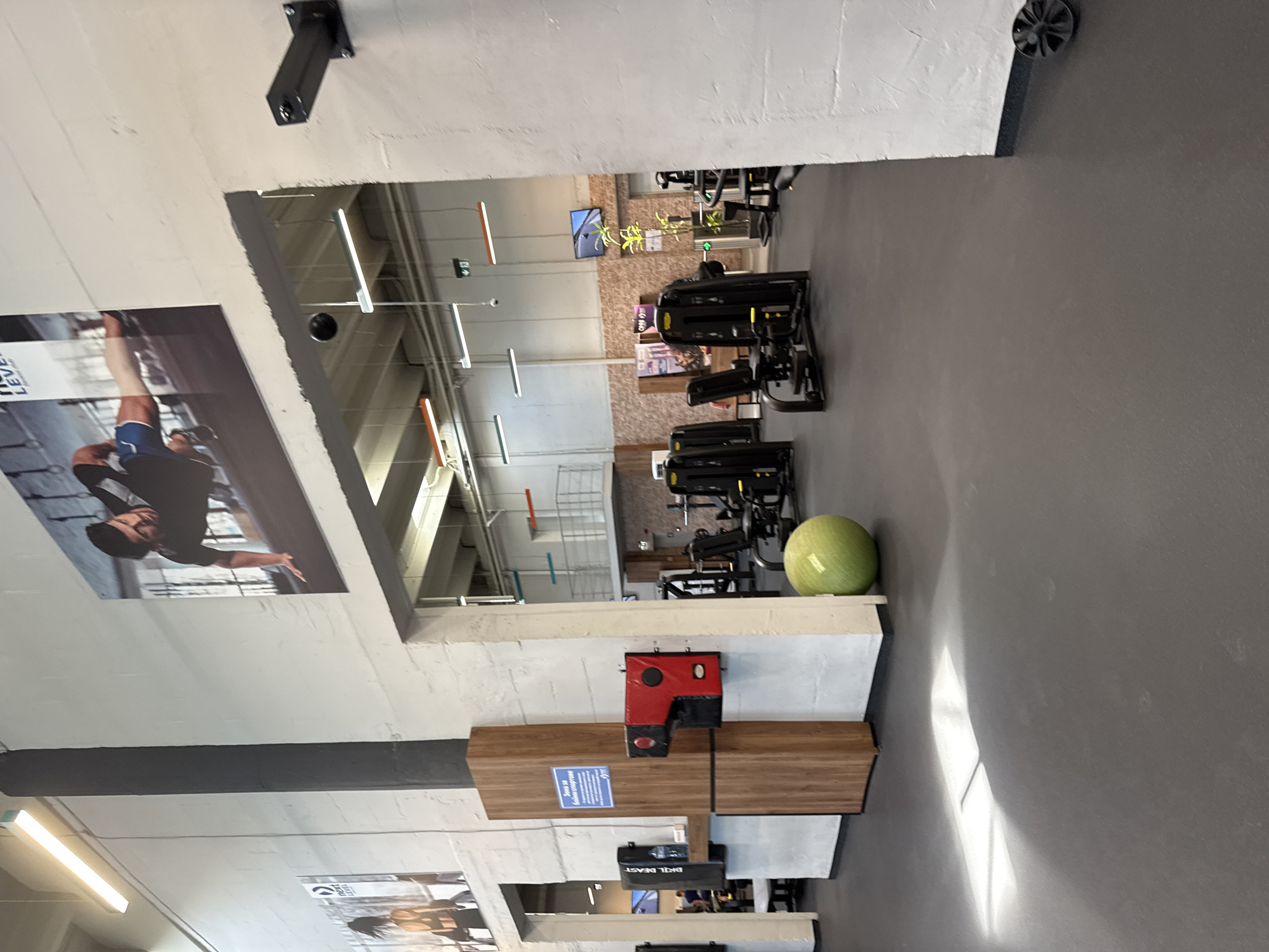 The image depicts the interior of a gym in Sofia, Bulgaria. Exercise machines are arranged on a gray floor. A green exercise ball sits near a shelf with a red and black object. A wooden structure with a blue sign is attached to the wall. Posters are mounted on the wall, one showing a man in athletic gear. Overhead, fluorescent lights are visible. The view is slightly distorted, possibly due to the camera's position or perspective.