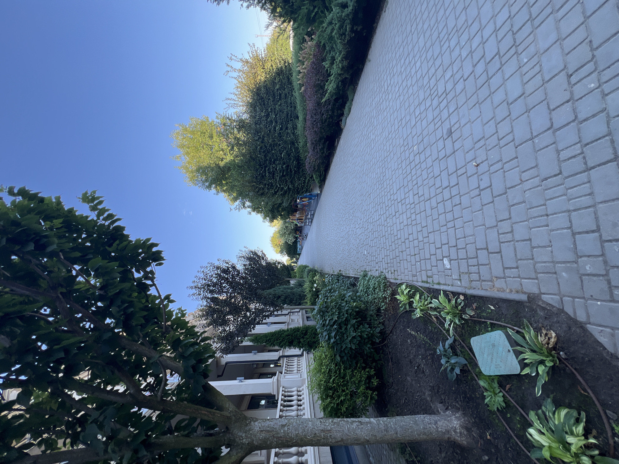 The image captures a paved walkway lined with manicured greenery and a building with classical architectural elements. The walkway, made of interlocking grey pavers, stretches into the distance, leading towards trees and shrubs. On the left, a building with white columns and a balustrade is visible, bordered by dense green bushes and plants. A large tree stands in the foreground, its trunk to the left. In the background, more trees and a bright blue sky complete the scene. The location is identified as Fontanka, Ukraine.
