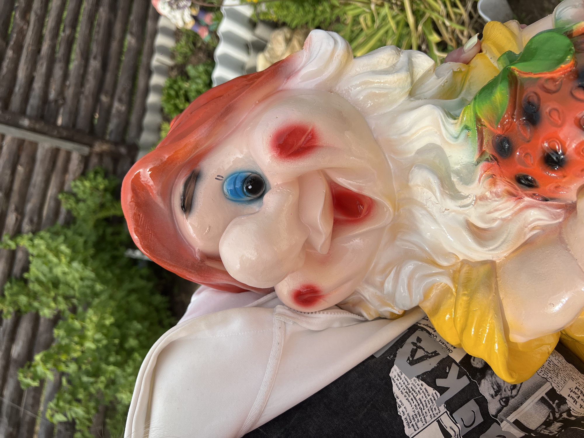The image displays a close-up, upward-tilted view of a vibrantly colored garden gnome or similar whimsical figurine. The figure has a pale, smooth complexion with prominent red blush spots on its cheeks and a wide, open-mouthed smile with red lips. A single blue eye with a black pupil is visible, accented by dark, simple lines for the brow or lashes. It sports bright red, sculpted hair, and a voluminous white, wavy element that resembles whipped cream or meringue, possibly serving as hair or a hat. This white feature is interspersed with yellow elements, possibly representing flowers, and green leaves. To the right, a large red, textured object resembling a strawberry or raspberry, complete with black seeds, is prominent.
The background is an outdoor setting, blurred but discernable, featuring a vertical wooden slat fence and lush green foliage. A metallic, corrugated surface is also partly visible in the upper background. Beneath the figurine, a white fabric item, possibly clothing or a covering, is present. Below this, fragments of black-and-white newspaper print can be seen, with partially legible text such as "Officers C...", "K", "U", "art," and "Earl's". The bright lighting suggests it is daytime. The scene is static, focusing solely on the details of the figurine and its immediate surroundings. The overall impression of the figure is joyful and caricatured. The location context is Chaikovsky, Russia.
