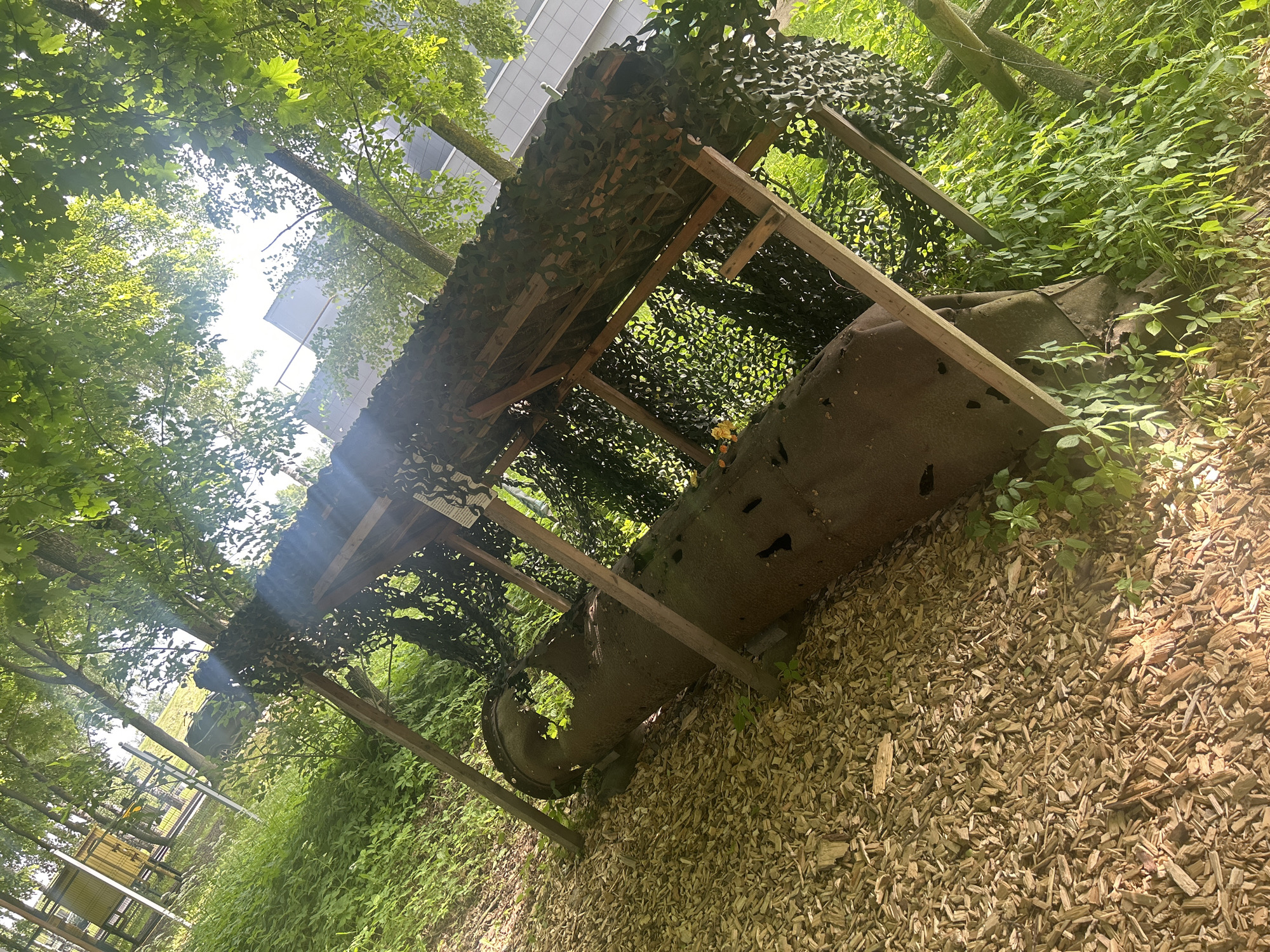 The image shows an outdoor scene with a large, rusted, cylindrical object lying on its side, partially covered by a wooden structure with camouflage netting. The object appears to be a piece of industrial pipe or a large artillery shell. The ground is covered with wood chips, and lush green vegetation surrounds the structure and the object. Trees with dense green foliage create a canopy overhead, with sunlight filtering through the leaves. In the background, a modern building with white walls is visible through the trees. The scene is likely part of an outdoor exhibit or a historical display. The location is identified as being in Shlisselburg, Russia. There are no people visible in the image.