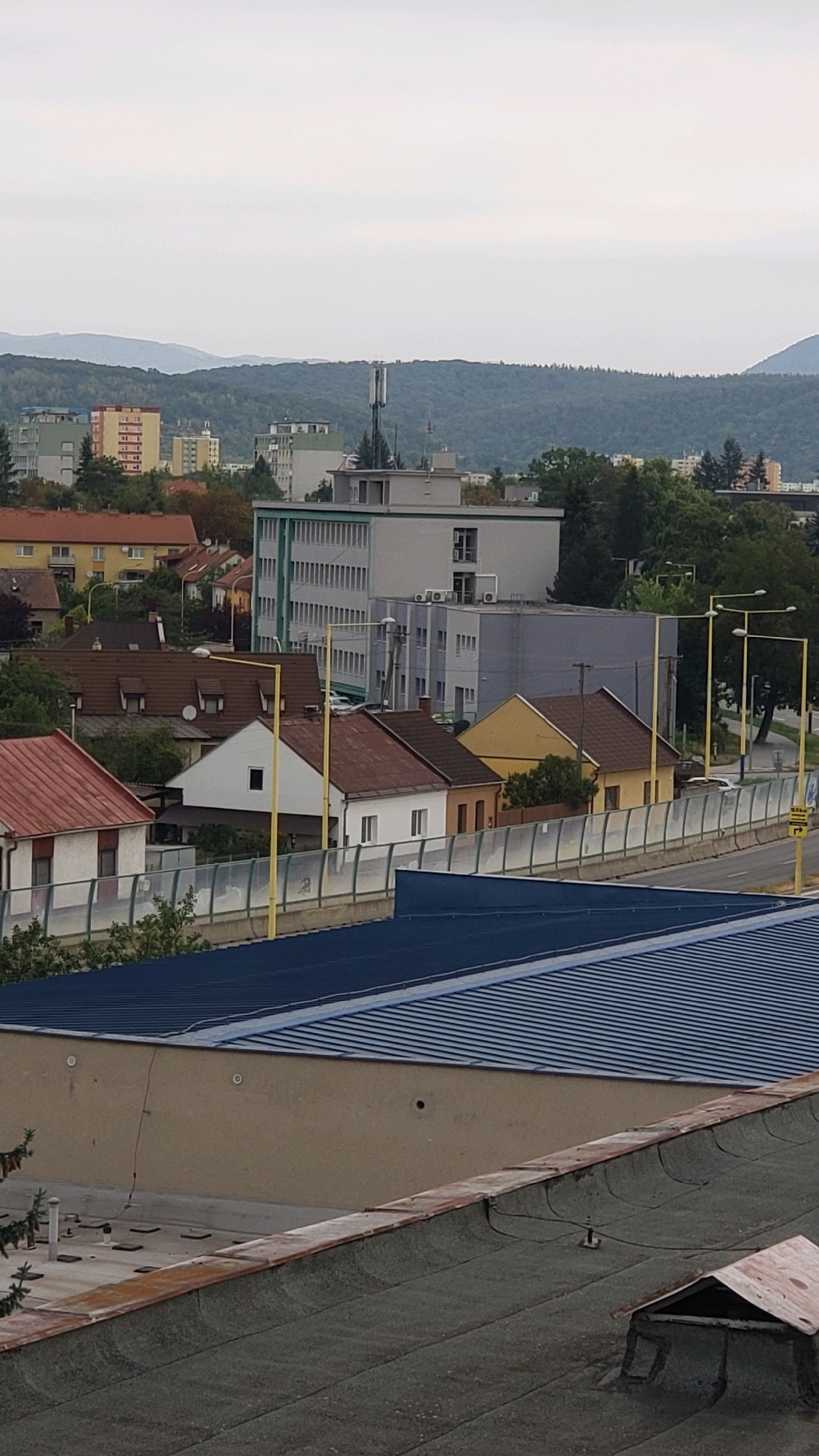 The image is an outdoor, mid-range shot of a town or city with buildings and hills in the background. In the foreground, there is a sloped rooftop with some rooftop equipment. Behind this, a light brown building is visible, with a blue corrugated metal roof. Beyond that, a row of residential buildings with red and white facades and dark red roofs are lined up along a road. Some of these houses have visible chimneys. A fence with glass panels separates the road from the houses. Further in the background, a cell tower and several taller buildings, some with light-colored facades and many windows, are visible. The background consists of green, tree-covered hills under an overcast sky. The overall scene depicts an urban or suburban environment. The presence of a cell tower and the architectural style of the buildings suggest this location is in Eastern Europe, potentially Prešov, Slovakia, given its geographical context of hills and urban development.