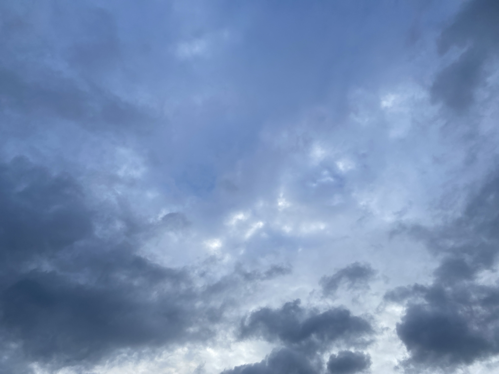 The image presents an expanse of the sky over Chaikovsky, Russia, dominated by an overcast to partially overcast cloudscape. Cumuliform clouds, ranging from light bluish-grey to dark grey, are distributed across the frame. Denser, darker cloud formations are interspersed with lighter, more diffuse areas where the sky appears brighter, suggesting varied cloud thickness and light penetration. No other subjects, objects, individuals, or actions are depicted in the scene.