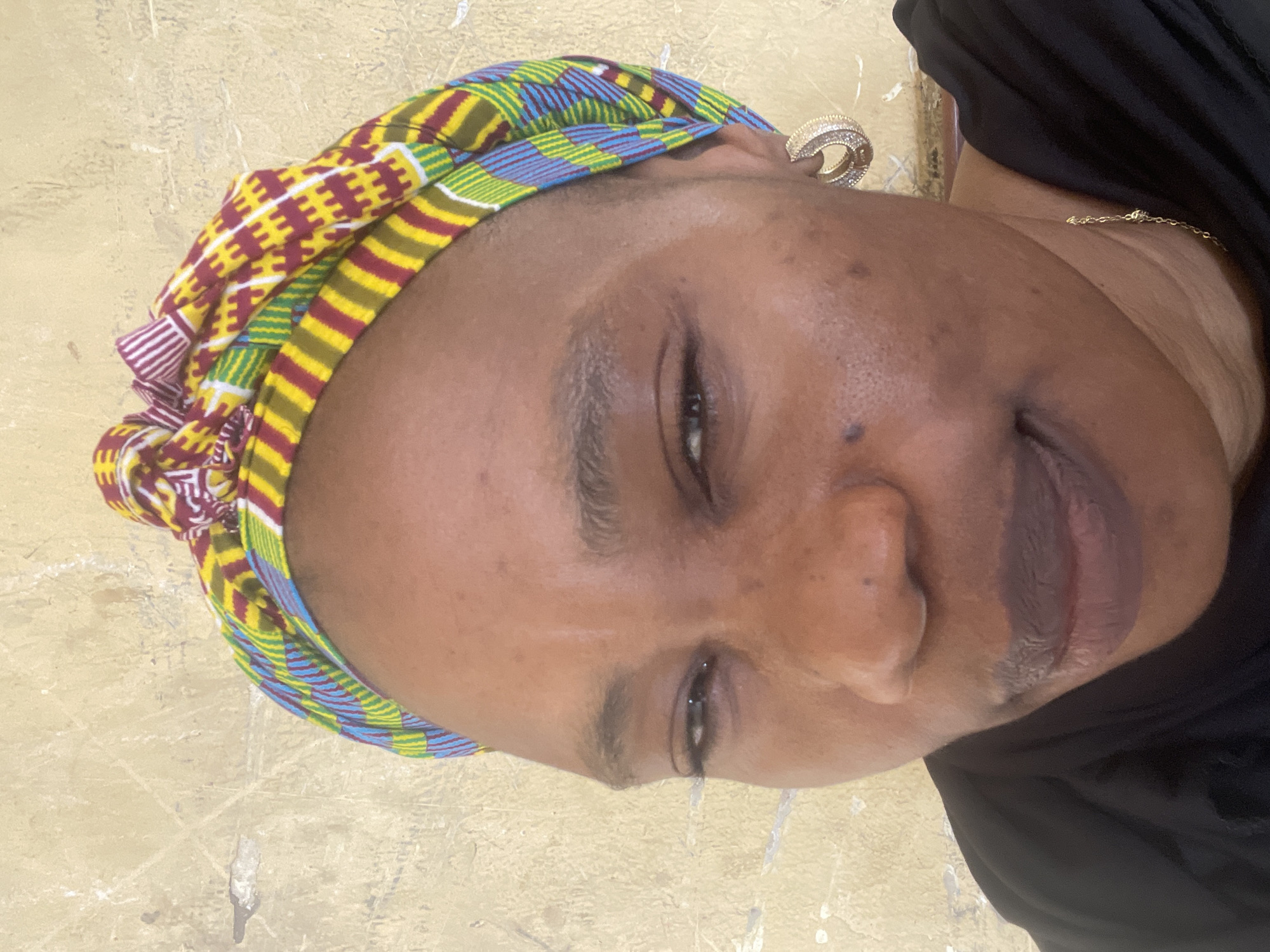 A close-up, upright portrait captures an adult individual, likely female, with dark skin, in Unguwar Rimi, Nigeria. The subject looks towards the viewer with a subtle, serene expression, showing a hint of a smile. They are adorned in a vibrant, multi-patterned headwrap, featuring rich hues of yellow, red, green, and blue in geometric designs and stripes, akin to Kente cloth. A prominent, gold-colored hoop earring, embellished with small gems, is visible on their right ear. A delicate gold chain necklace rests against their neck, peeking from beneath a plain black garment. The background consists of a simple, light-colored, textured wall. The lighting is soft and natural, suggesting the photograph was taken during daytime hours. There is no visible text in the image.