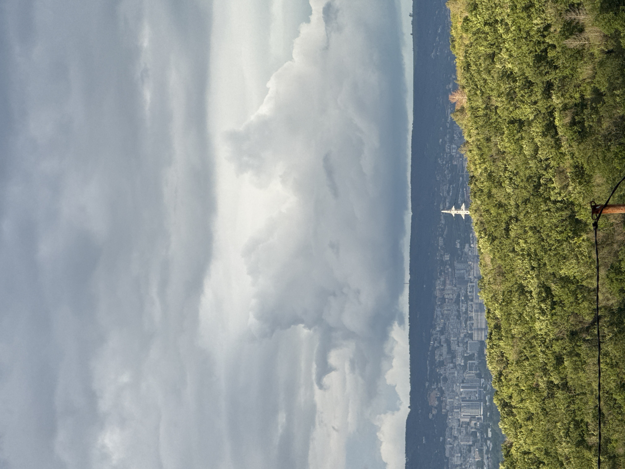 The image depicts a cityscape situated between a mountain range and lush green vegetation. The sky is overcast with varying shades of gray clouds. A transmission tower stands near the forested area. The city appears dense with buildings. The image appears to have been taken in a location overlooking Konstantinovo, Bulgaria, though this cannot be definitively confirmed without additional contextual clues.