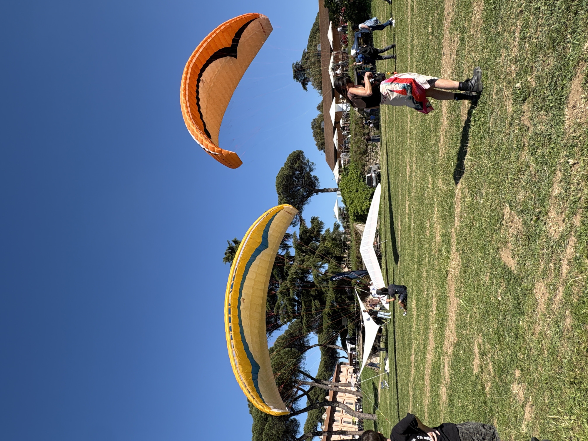 The image depicts two paragliders over Rome, Italy on a sunny day. The paraglider in the foreground is yellow with blue trim, while the one in the background is orange with black trim. Below the paragliders, people are standing on a grassy field. A hang glider is positioned on the ground nearby. In the background, buildings and trees can be seen. A person in shorts and a dark shirt is standing to the right of the hang glider and appears to be taking a photo.