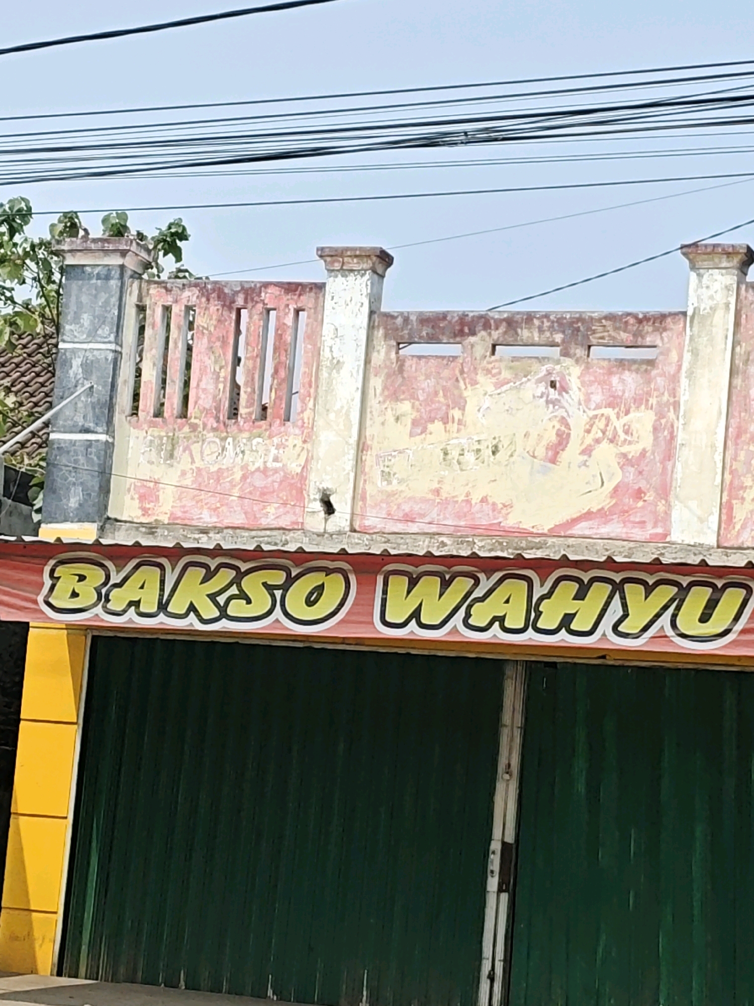 A storefront with a red banner displaying "Bakso Wahyu" in yellow lettering is visible, set against a backdrop of a weathered, pink and cream-colored building with ornate pillars. The building's facade is peeling and shows signs of age, with some visible graffiti or faded lettering. Above the building, a tangle of electrical wires crisscrosses the sky. The storefront itself features a dark green, corrugated metal roll-up door, with a yellow vertical support on the left side. The image appears to be taken in Kemuning, Indonesia, given the context of the requested location.