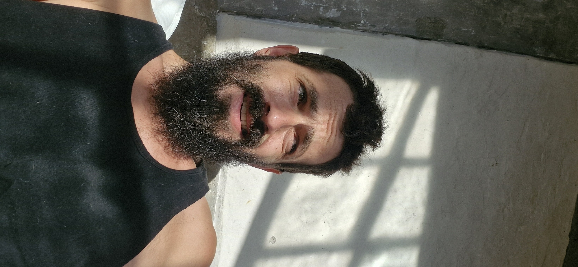 A man with dark hair, a beard, and a black tank top is photographed from the chest up. He is looking upwards and to the right, with a slight smile. The background is a wall with shadows cast on it, possibly from a window or fence. The lighting suggests it is daytime. The setting appears to be outdoors.
