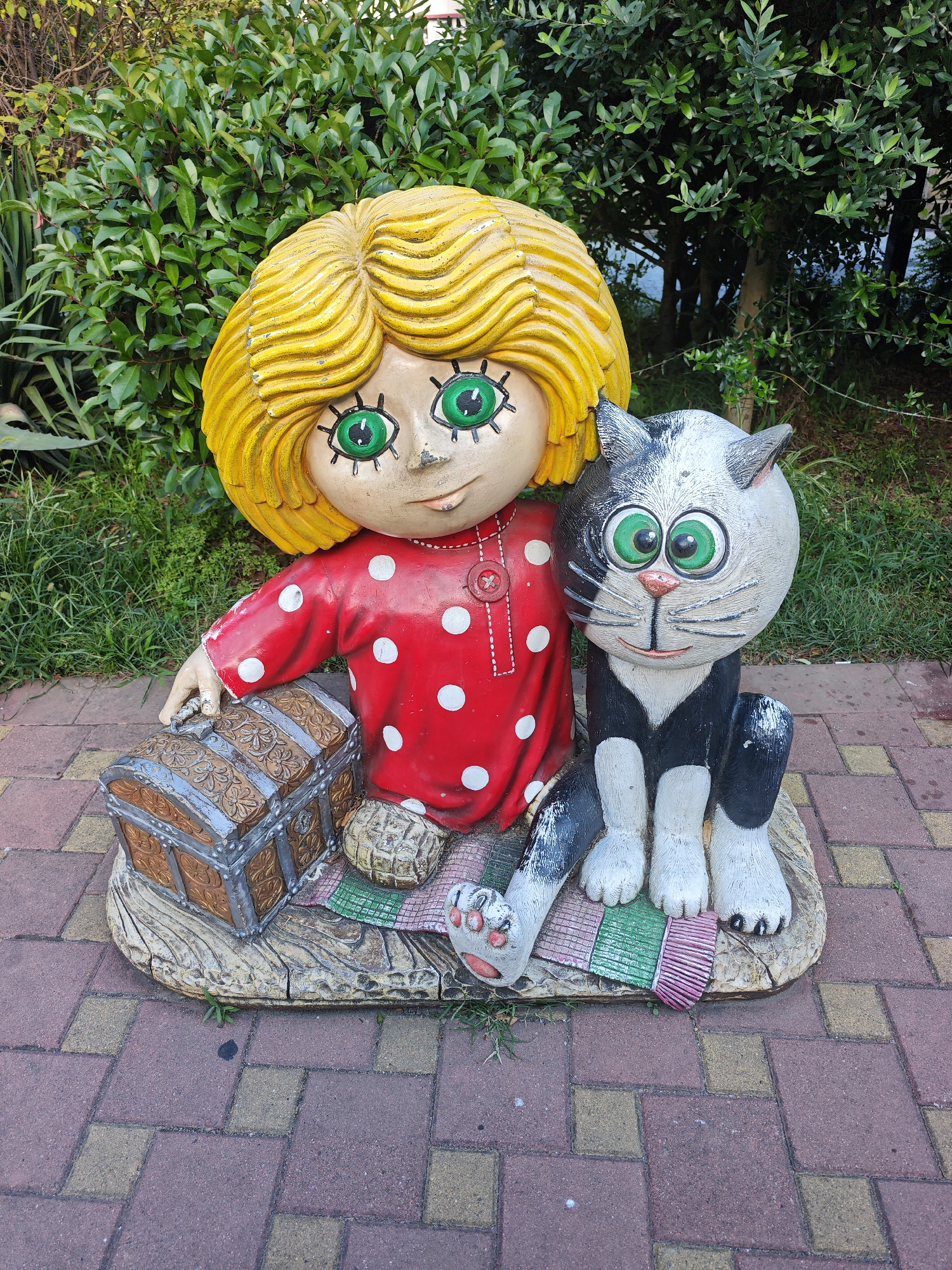 This image depicts a whimsical garden sculpture of a child and a cat sitting together on a decorative base. The child has bright yellow, wavy hair and large, green eyes. She is wearing a red dress with white polka dots. Next to her sits a black and white cat, also with large green eyes and a friendly expression. In front of the child is a small, ornate treasure chest. The sculpture is placed on a patterned brick pathway, with lush green foliage in the background. This scene is located in the Dendrarium Botanical Garden in Sochi, Russia, a popular tourist destination known for its diverse plant collections and decorative garden features.