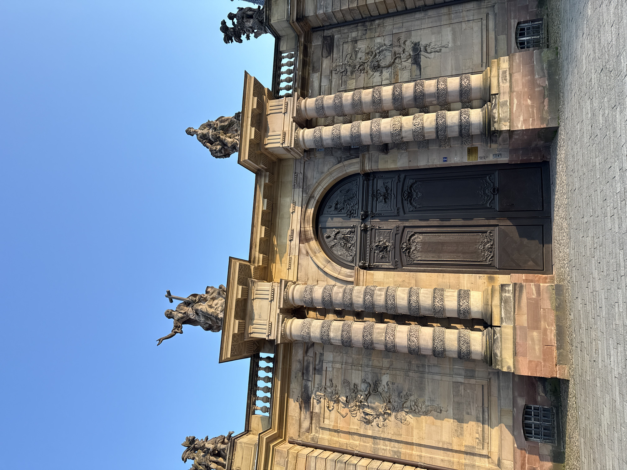 The image shows a partial view of the Saint-Pierre-le-Jeune Protestant church in Strasbourg, France. The focus is on a section of the facade featuring a large, ornate wooden door flanked by pairs of decorated columns. Above the door, there are architectural embellishments and sculptural groups, with additional sculptures positioned at the top corners of the building. The sky is clear and blue in the background. A small section of cobblestone paving is visible in the lower right corner of the image.