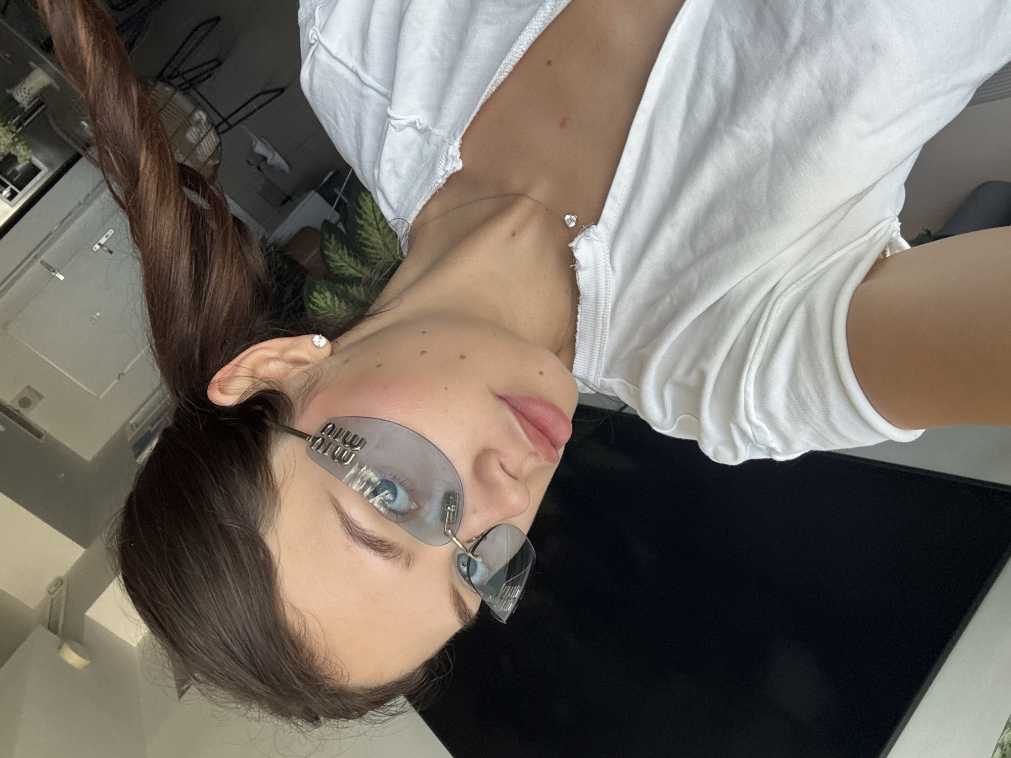 A young woman with dark brown hair is shown in a close-up shot, with her head tilted to the side. She is wearing a white t-shirt, a delicate silver necklace with a single diamond pendant, and distinctive clear-rimmed sunglasses with a heart-shaped lens and a coiled wire detail at the bridge. Her eyes, a striking blue, are visible through the lenses. She has a few freckles on her cheeks and appears to be wearing light makeup. The lighting suggests it might be daytime, possibly indoors with natural light filtering in. The background is mostly out of focus, but hints of a room with furniture and plants can be discerned. There is no discernible text in the image.