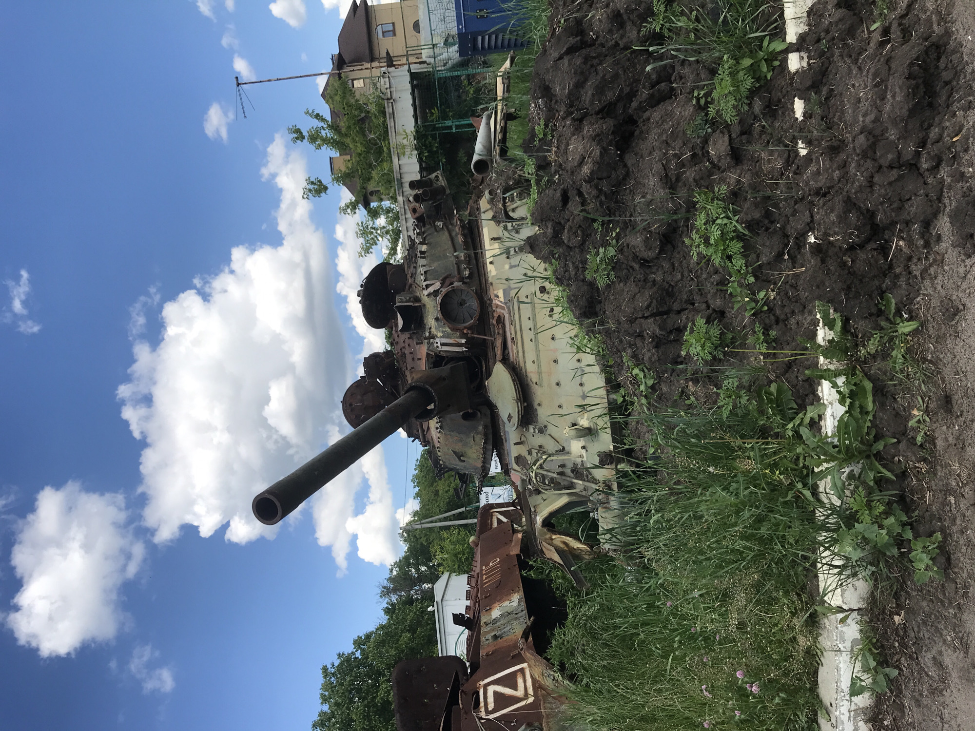A photograph, rotated ninety degrees counter-clockwise, depicts a heavily damaged and rusted military vehicle, identified as an MT-LB (Multi-purpose light armored towing vehicle), resting outdoors under a partly cloudy blue sky. The vehicle's main gun barrel points towards the upper left corner of the frame (when correctly oriented). Its exterior is extensively scarred, with visible rust and missing components, suggesting it has been destroyed and abandoned.
A prominent white letter "Z" with a black outline, a symbol associated with Russian military forces, is painted on a rusted section of the vehicle's lower hull. Above the "Z", the Cyrillic letters "МТЛБ" are visible, confirming its identification.
The vehicle is positioned next to a large pile of dark, disturbed earth, partially covered with green grass and weeds. More vegetation, including tall grass, surrounds the vehicle on the ground. In the background, residential buildings with brown roofs are visible, along with green trees and a tall antenna structure. The bright daylight and green foliage indicate the image was taken during spring or summer, likely in the daytime. No people are present, and no activity is occurring, capturing a scene of post-conflict destruction and abandonment.