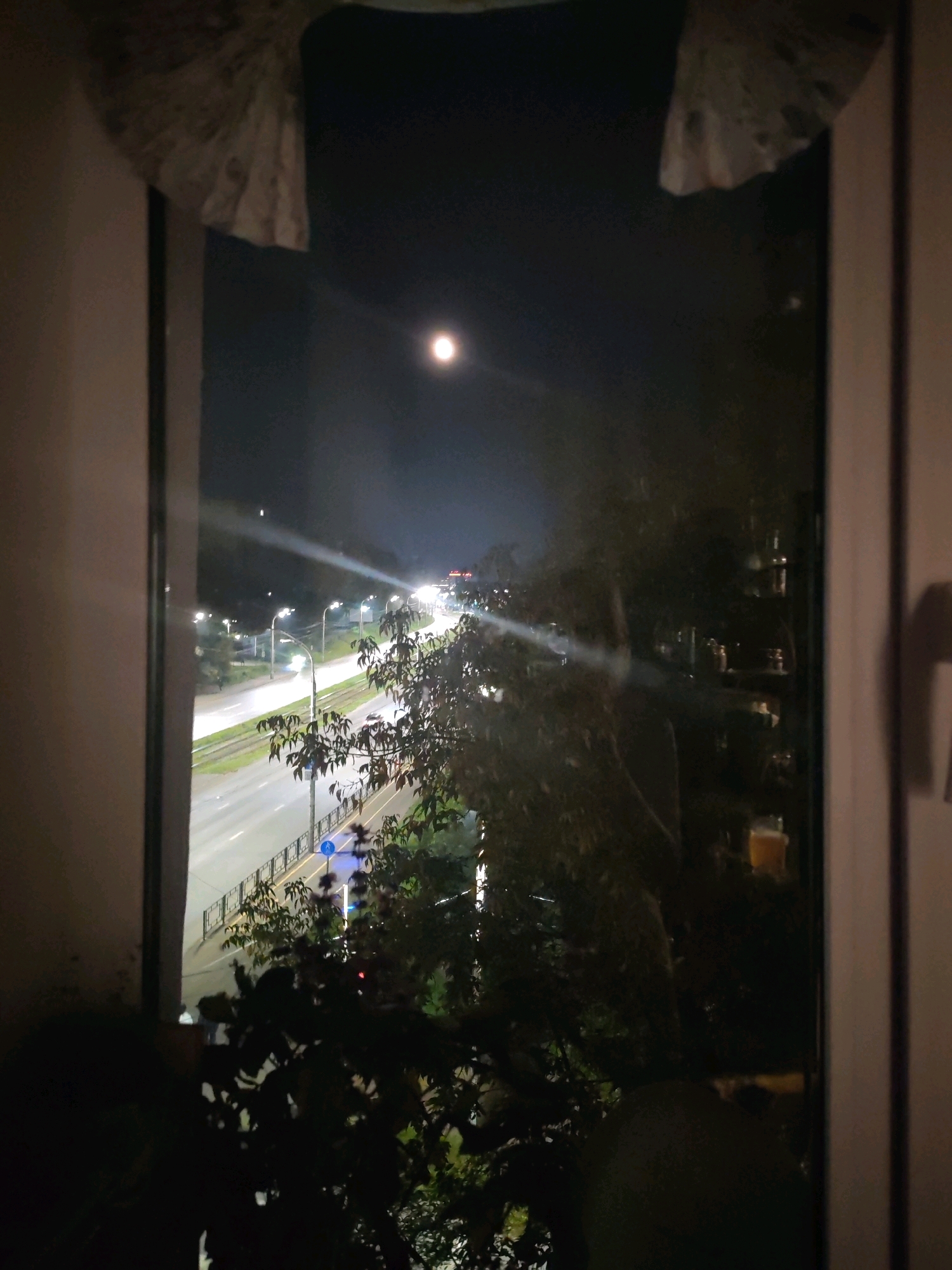 This is a nighttime, outdoor, medium shot taken from a window overlooking a street with moderate traffic. The view is partially obstructed by trees in the foreground and curtains on the sides. A full moon is visible in the dark sky. The street is well-lit by streetlights, and there are reflections on the wet asphalt. The scene is likely from Izhevsk, Russia, based on the prompt.