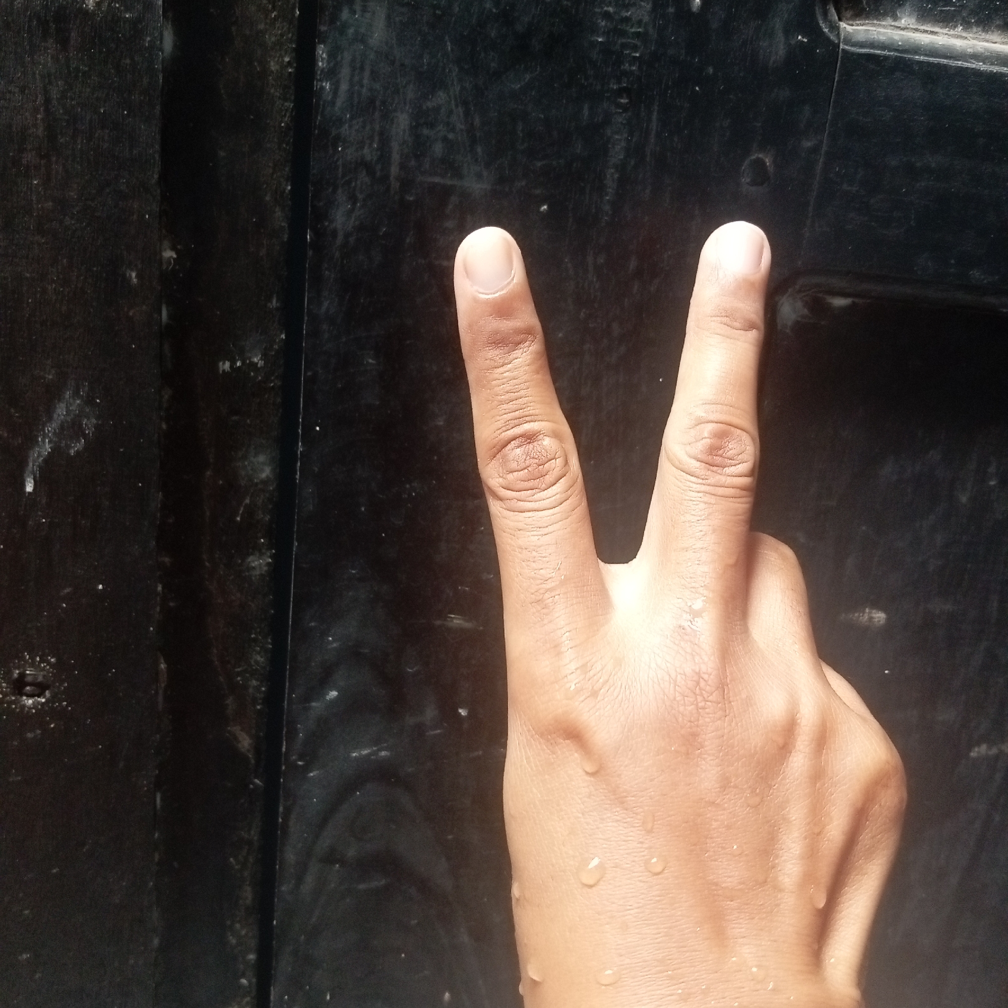 A close-up shot shows a person's hand, with two fingers extended in a V-shape, against a dark, textured background. The hand appears to be damp, with several small water droplets visible on the skin. The background is likely a dark, wooden surface, possibly a door or a wall, with visible grain and some smudges or imperfections. The lighting is somewhat dim, casting subtle shadows on the hand and highlighting the texture of the background. The specific location in Kediri City, Indonesia, cannot be determined from the visual information provided.