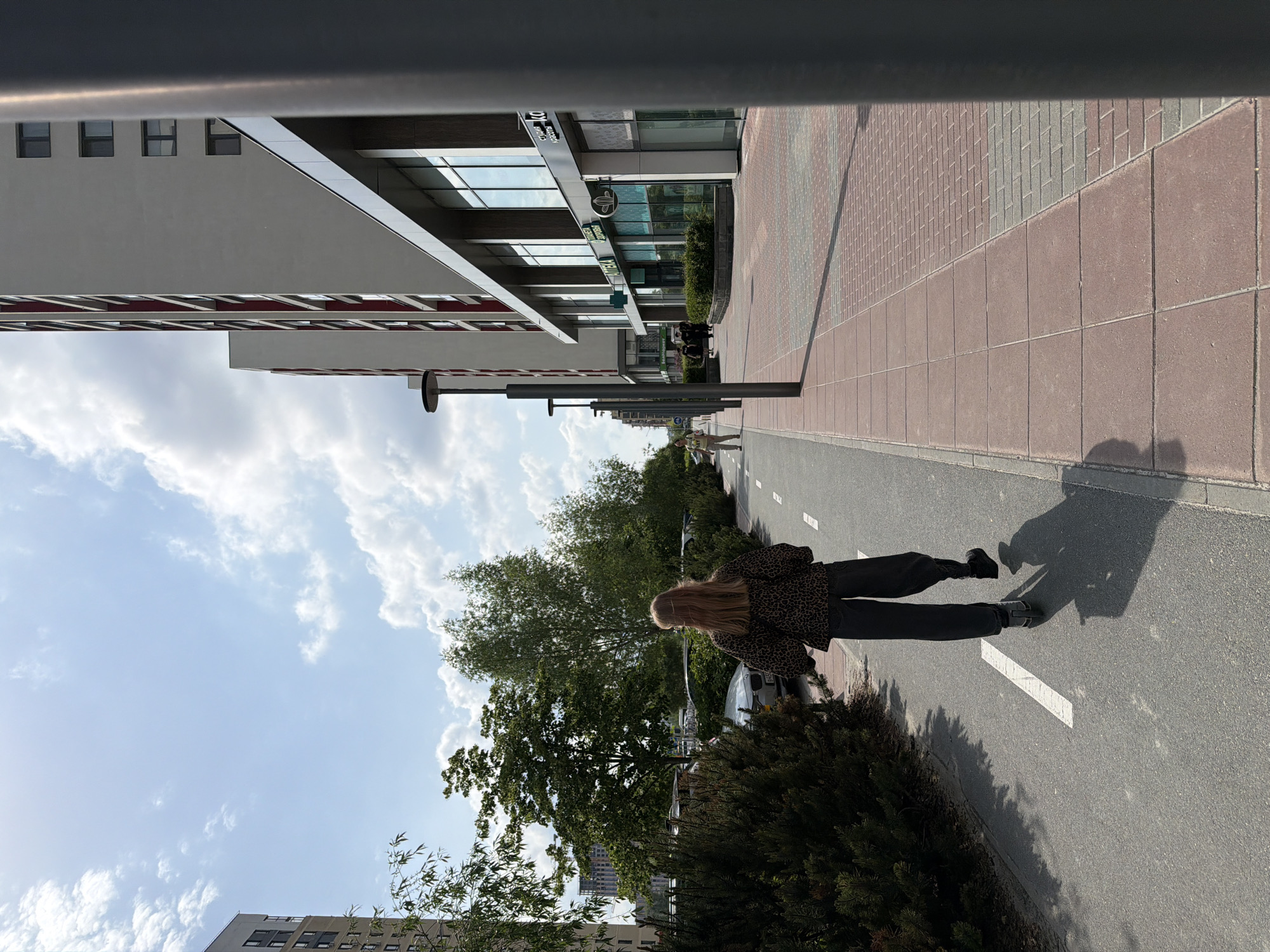 A person is walking on a paved bike path in an urban environment, parallel to a sidewalk with brick tiles. The bike path has a white line marking. To the left is a row of bushes and trees. In the background are buildings with glass facades and grey cladding, visible partially in the sky. Street lamps are spaced along the sidewalk. The scene is in Tyumen, Russia, based on the surrounding architecture and streetscape.