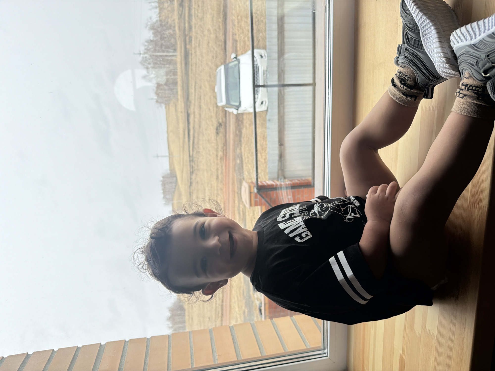 A young child wearing a black "Game Station" t-shirt and zebra-print socks is sitting on a wooden windowsill with legs extended. The child is wearing gray athletic shoes. The background shows a view outside a window. A white vehicle is parked in a yard with a partly cloudy sky. The photograph was taken in Krasnoyarsk, Russia, based on the building style and landscape.