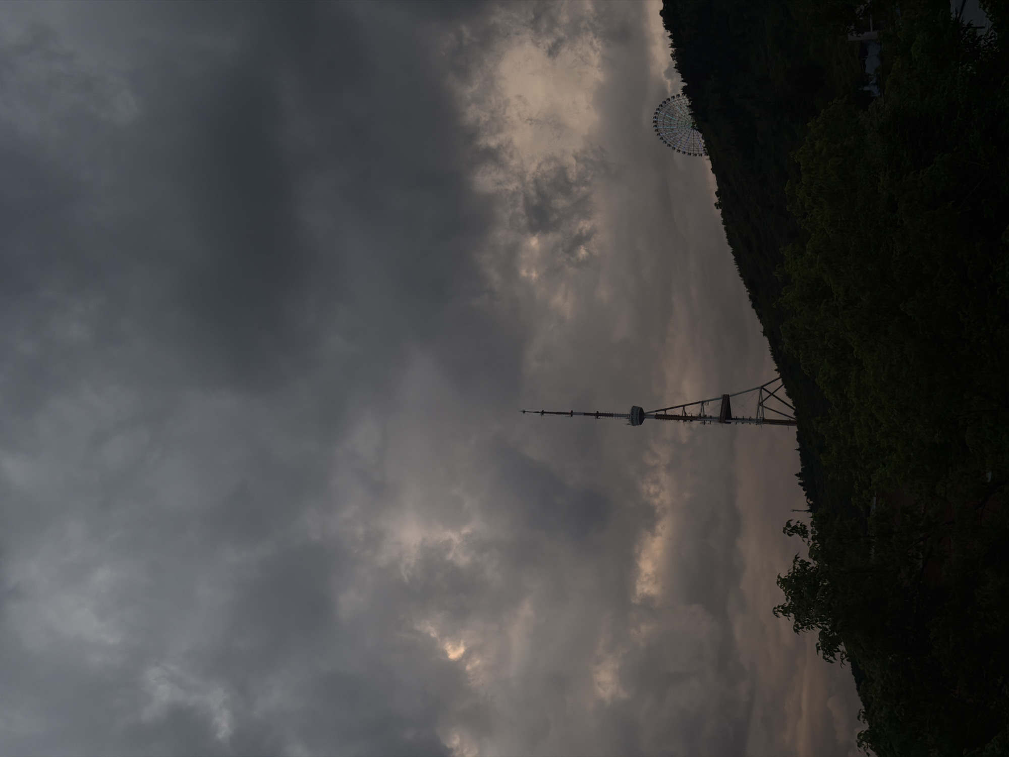 The image shows a view of the Tbilisi TV Broadcasting Tower in Tbilisi, Georgia, under a cloudy, overcast sky. The tower is silhouetted against the sky, and its structure is partially obscured by clouds. A ferris wheel is barely visible in the upper right corner. Dense vegetation covers a hillside to the right of the tower.