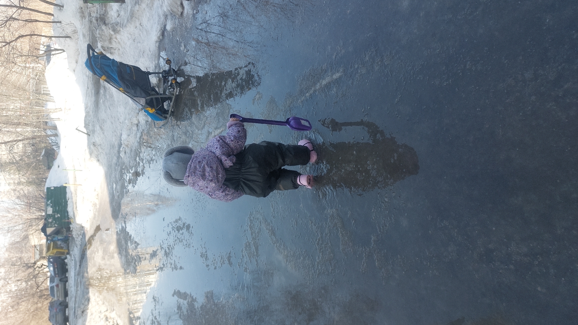 A young child in a lavender floral jacket, dark trousers, pink boots, and a gray hat stands in a puddle on a paved surface. The child holds a purple shovel. To the left, partially obscured by snow, is a blue and gray stroller. Beyond the stroller, there are bare trees and buildings. Cars are parked along the side of the street. The location is Novosibirsk, Russia.
