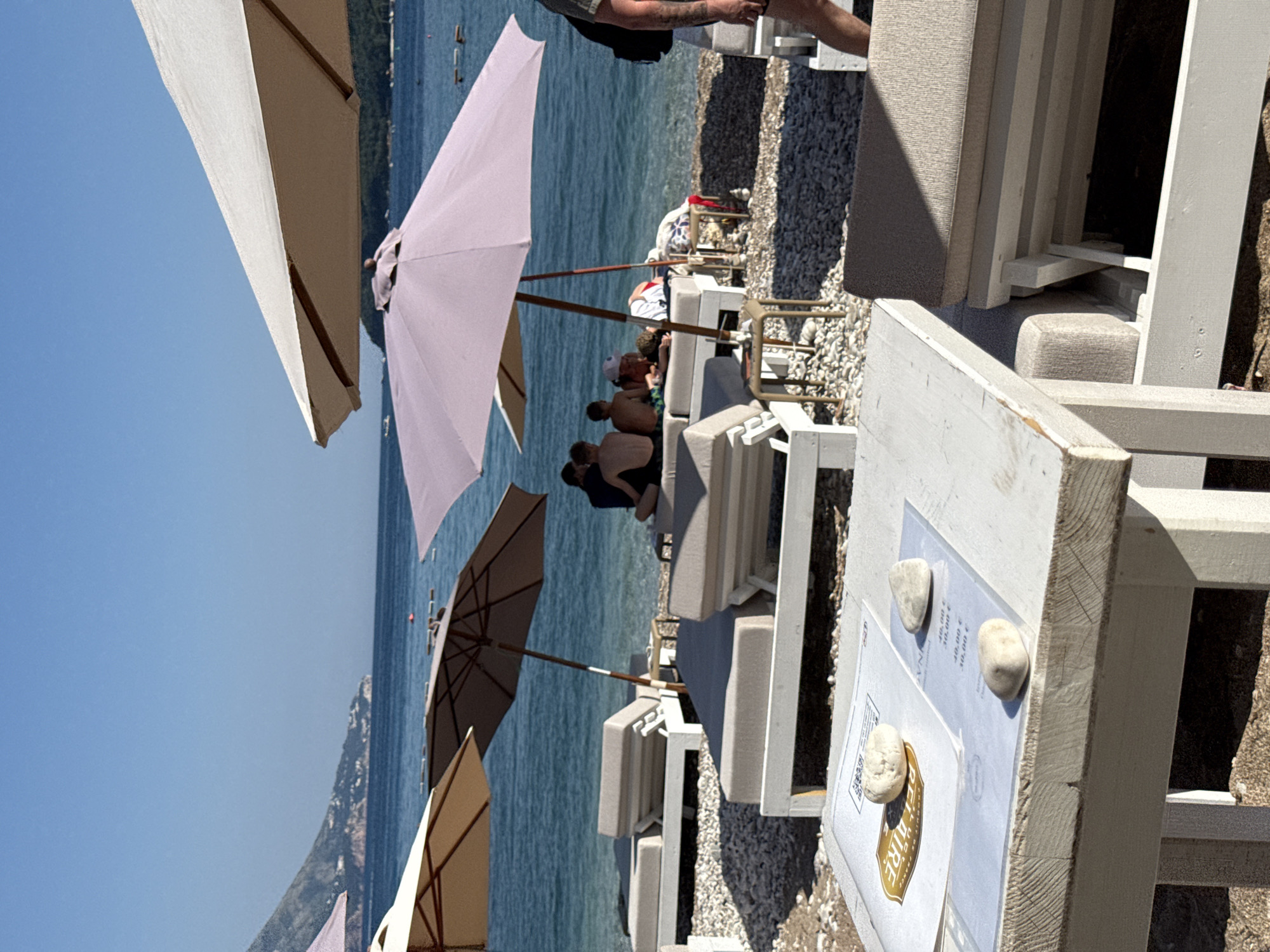 The image depicts a sunny day on a pebbly beach in Budva, Montenegro, near the Adriatic Sea. The foreground shows a white wooden table with a menu, secured by three small, light-colored stones. Beyond the table, several white lounge chairs with grey cushions are arranged, some occupied by people. In the middle ground, under a large lilac umbrella, a group of individuals are seated on lounge chairs, appearing relaxed. To the left, another umbrella provides shade over more lounge chairs. The turquoise sea stretches to the horizon, where a hilly coastline with sparse vegetation and buildings is visible. The sky is clear and blue. A person in the right foreground, partially visible from the waist down, is walking away from the camera. The overall scene conveys a peaceful beach resort atmosphere.