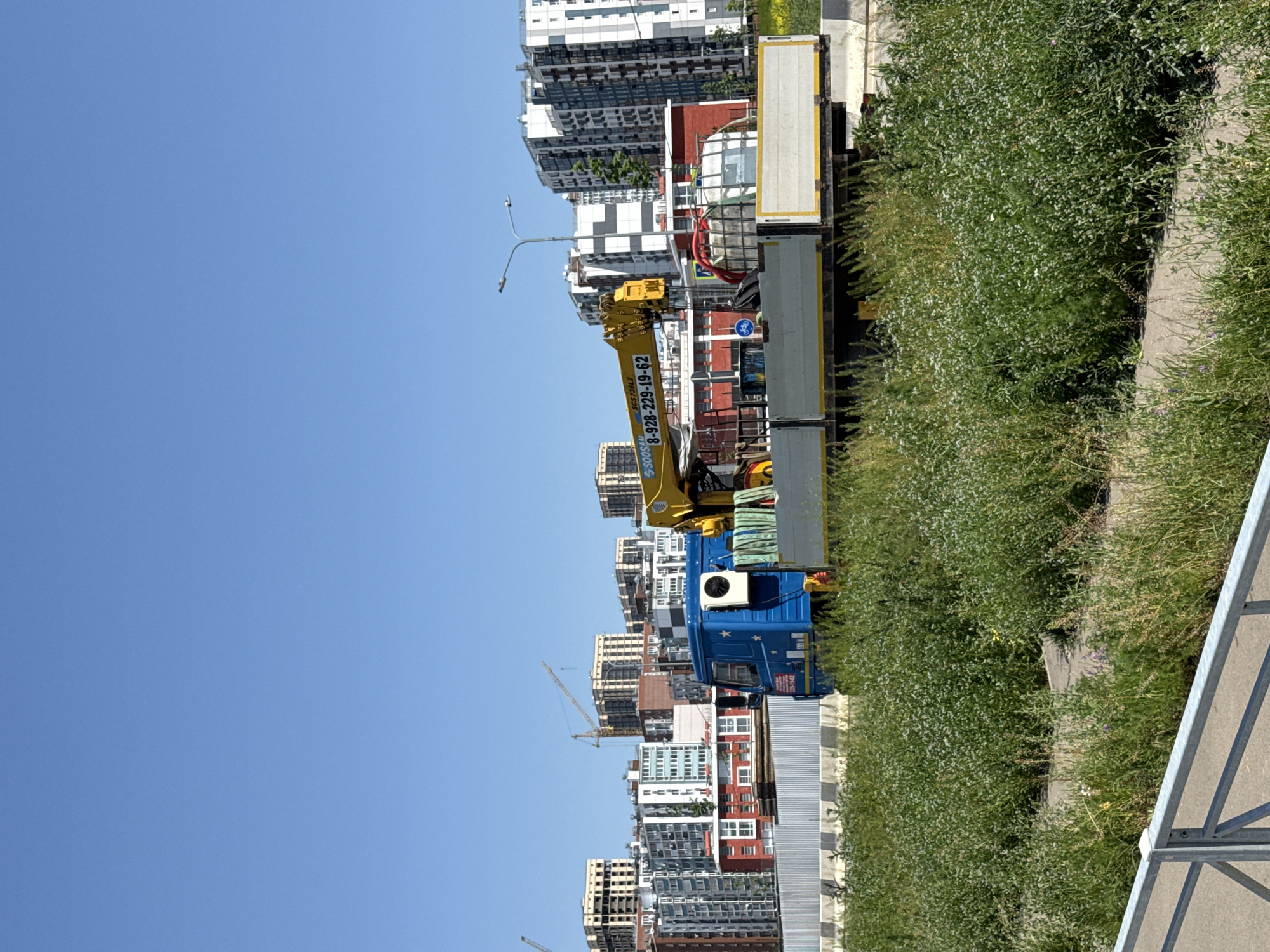 The image depicts an urban landscape in Rostov-on-Don, Russia. A blue and white truck with a yellow crane is parked alongside a road adjacent to a dense patch of green vegetation. The truck appears to be a work vehicle based on the crane attachment. Behind the truck, a series of high-rise apartment buildings dominate the skyline, reflecting a mix of residential architecture. The sky above is clear and blue.
