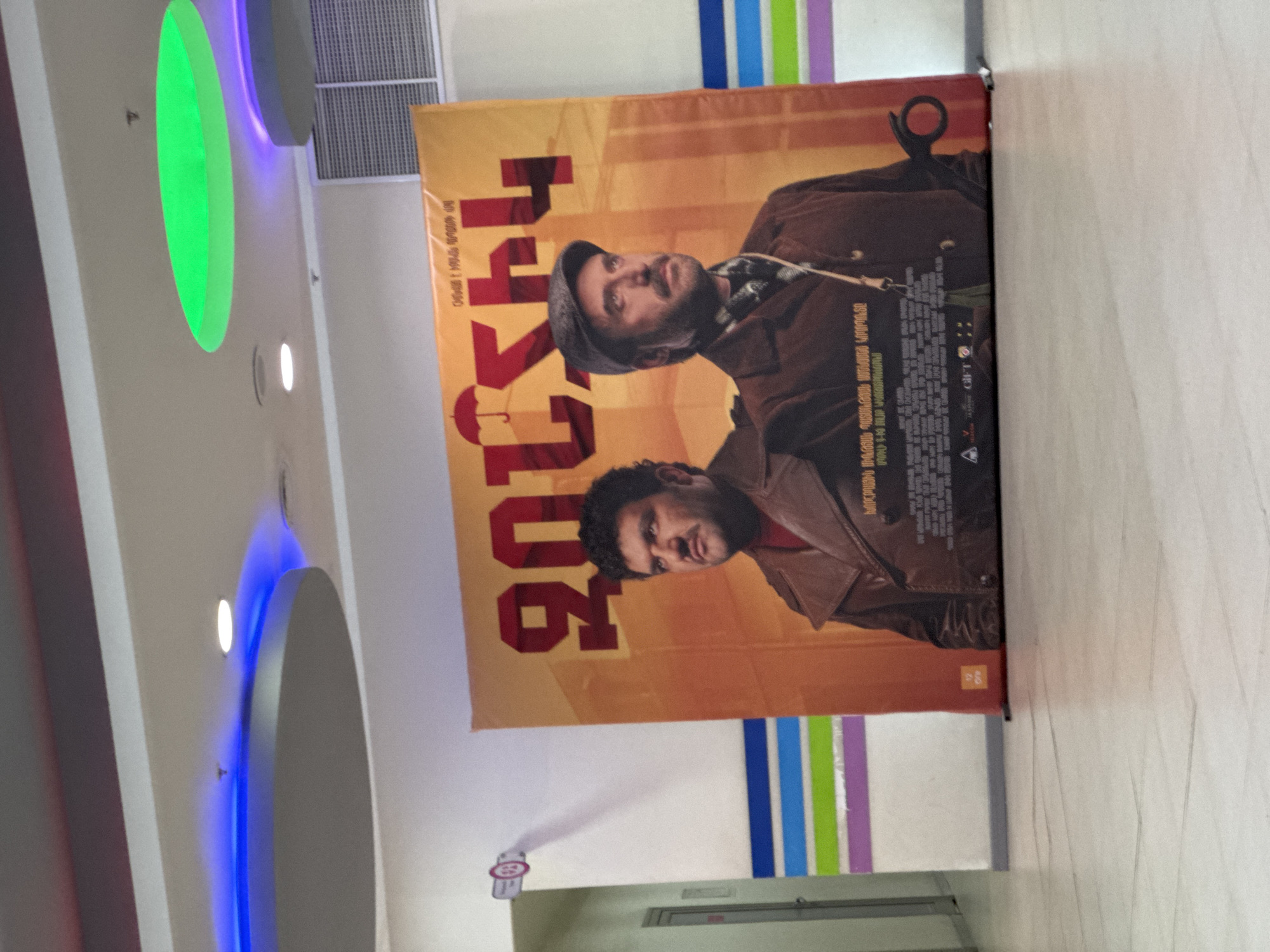 The image shows a movie poster on a wall indoors. The poster is for a film whose title is written in Georgian script, with the numerals "206" prominently displayed. Two men are featured on the poster, dressed in what appears to be older style clothing. The wall has multi-colored vertical stripes. Below the poster, there's a low, horizontal structure, possibly part of the wall or a fixture. Above the poster, a vent is visible on the ceiling. There are also decorative lighting fixtures in the ceiling of the room, including a large green oval and a similar blue oval. This is located in the city of Yerevan, Armenia.