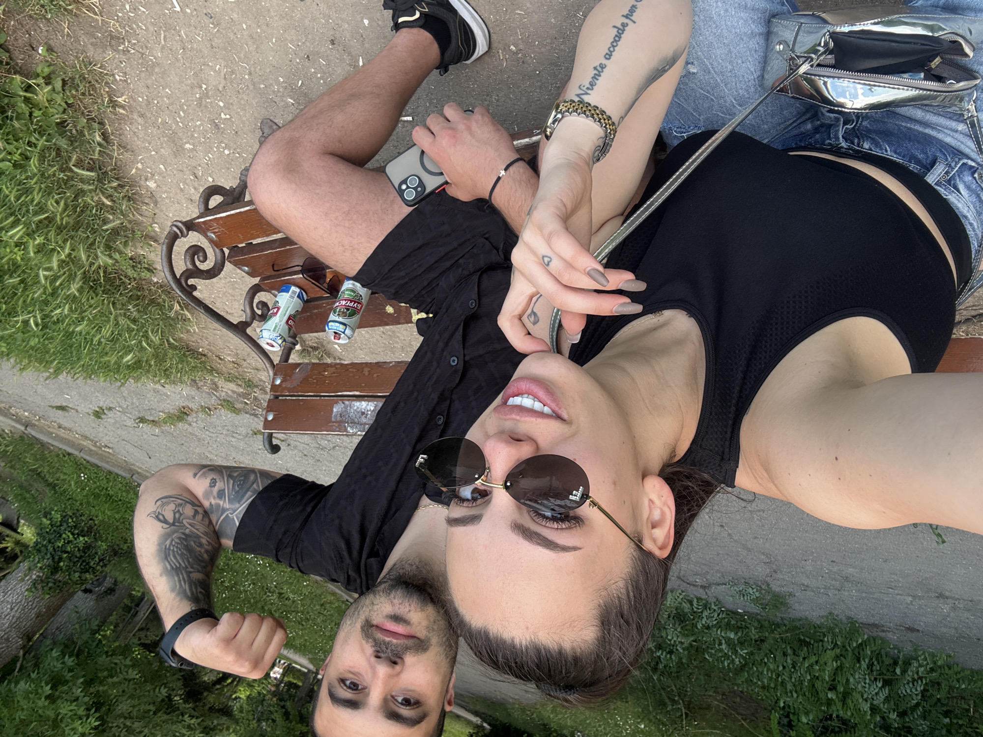 Two individuals, a man and a woman, are captured in a close-up, slightly tilted selfie-style image while relaxing on a park bench in Varna, Bulgaria. The setting is outdoors, featuring a paved ground, green grass, and visible trees and bushes, suggesting a park or public green space under bright, clear daylight.
The woman is positioned closer to the camera, wearing dark round sunglasses, a black crop top, and blue denim shorts. Her left forearm displays a tattoo reading "Niente accade per" and a small heart tattoo is visible on her hand, which is adorned with painted nails and a gold-colored watch. A silver metallic shoulder bag rests beside her.
The man is sitting next to her, wearing a black button-up shirt and dark shorts. His left arm is extensively tattooed with intricate designs, and he wears a black watch or bracelet. He holds a smartphone with a magnetic ring accessory. Both individuals are looking towards the camera with relaxed expressions.
On the wooden bench, next to the man, are two discarded beer cans. One prominently displays "EYPAOA" (Evropa) and "BECTHA" (Vestna/Vestnik) in Cyrillic, while the other green-labeled can partially shows what appears to be "ASTIKA," a Bulgarian beer brand. Another pair of sunglasses rests on the bench. Both subjects appear casual and at ease.