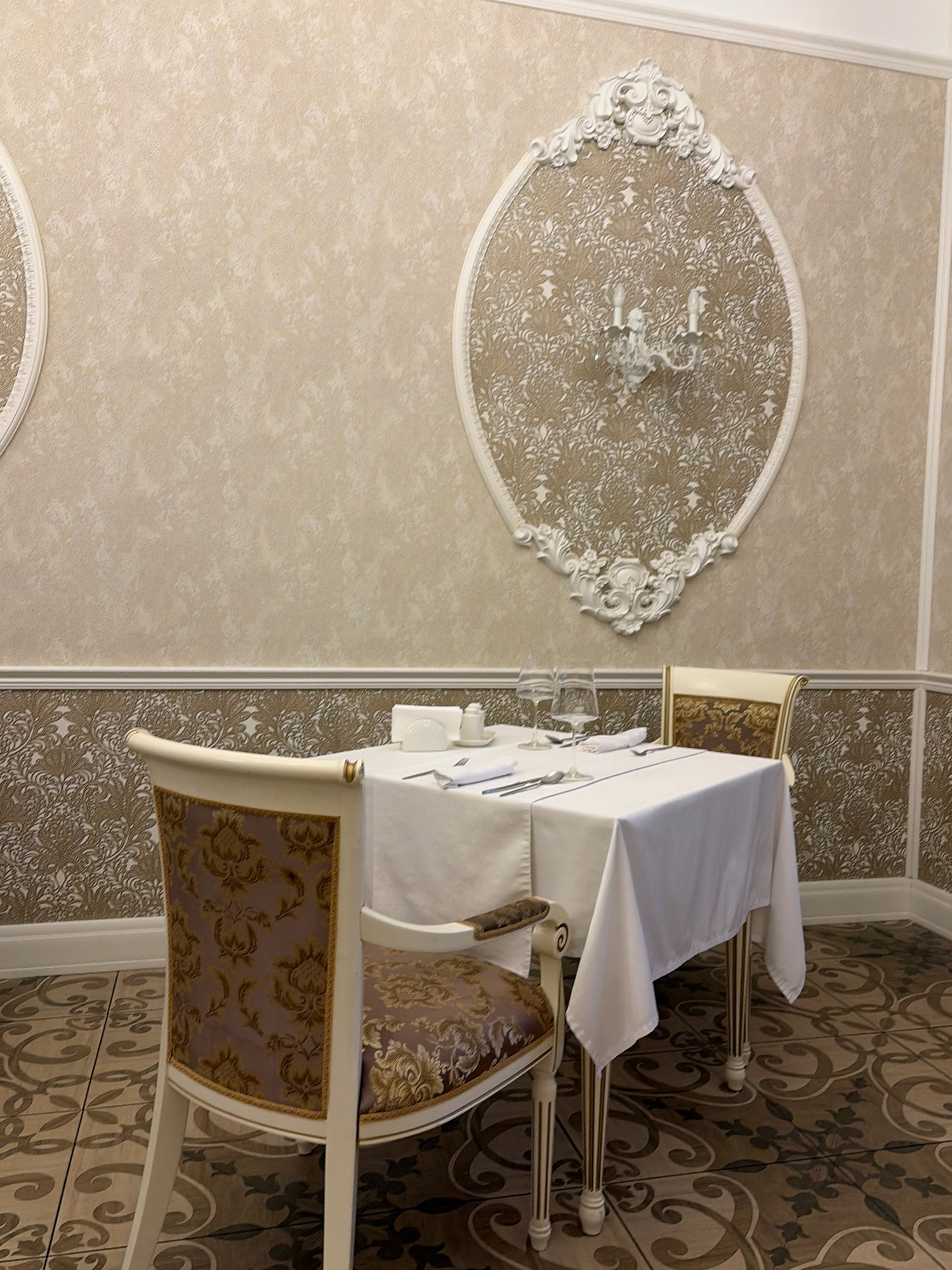 This image depicts a formal dining setting within what appears to be a restaurant or dining hall. The room is decorated with ornate wallpaper featuring a damask pattern in light and dark beige tones. A large, oval decorative frame with intricate white plasterwork is mounted on the wall, containing a textured wallpaper and a white candelabra.
In the foreground, a white-clothed dining table is set for two. Two wine glasses, cutlery, napkins, and salt and pepper shakers are visible on the table. Two chairs, upholstered in a patterned fabric of purple and gold, are positioned at the table. One chair is mostly visible on the left side of the frame, and the back of another chair is visible on the right. The floor is tiled with a brown and beige mosaic pattern.
The scene is static, with no people or visible activity. The lighting is soft, suggesting an indoor setting possibly during the day or evening. The overall impression is one of elegance and a classic, traditional aesthetic.
Regarding the location in Angarsk, Russia, there is no specific visual information in the image that can confirm or deny this location. The interior design elements are not unique to any particular geographical region or city.