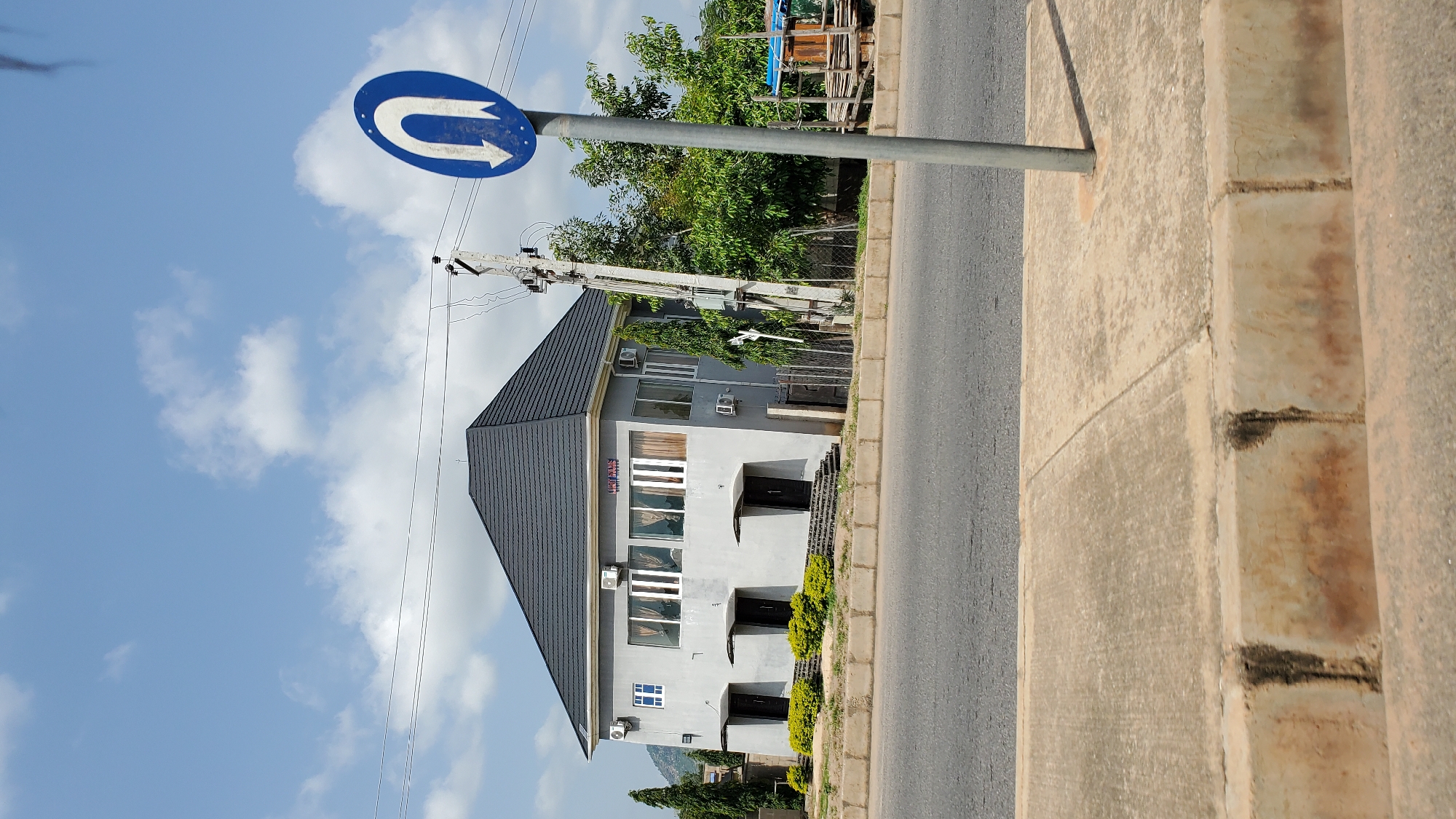 The image shows a two-story grey building with a dark grey pitched roof, located in Jalingo, Nigeria. The building has multiple windows on the upper floor, some with curtains, and three doors on the ground floor, each with a small canopy above it. There are air conditioning units mounted on the exterior walls. Green bushes are planted in front of the building, and a concrete pathway leads to the doors. To the right of the building, there is a U-turn traffic sign with a white arrow on a blue circular background, mounted on a metal pole. Electrical poles and wires are visible behind the building and to the right. Trees and vegetation are present in the background, along with a small blue-roofed structure and some wooden structures. A paved road runs in front of the building, and a concrete sidewalk is visible in the foreground. The sky is blue with scattered white clouds.