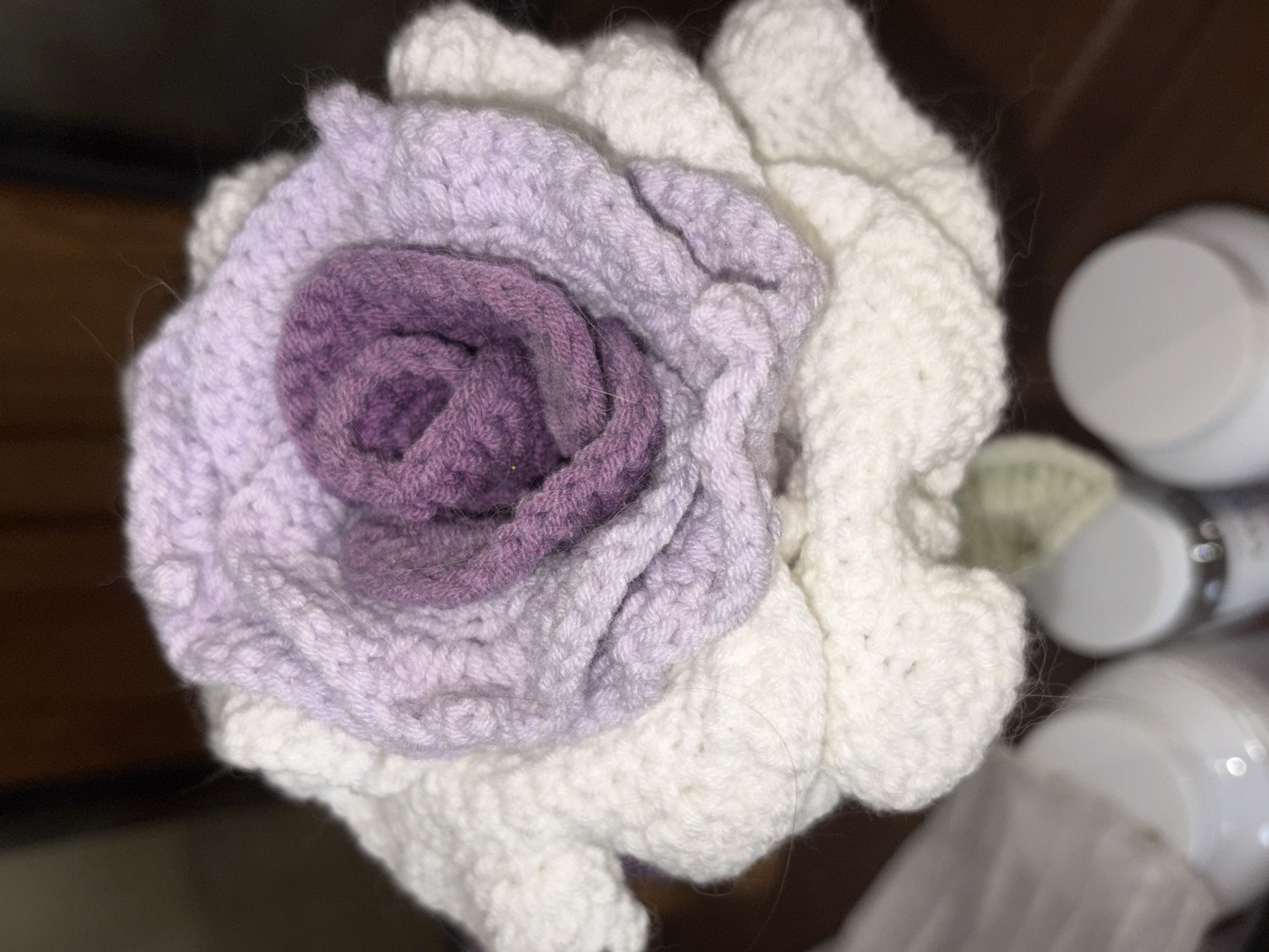 A close-up, slightly out-of-focus shot captures a hand-crocheted flower with multiple layers of petals. The innermost petals are a deep purple, transitioning to lighter shades of lavender and then to a prominent white outer layer. The texture of the yarn is clearly visible, with individual stitches forming the petal shapes. The flower appears to be made of wool or a similar soft fiber. In the lower portion of the frame, partially visible, are the tops of white cylindrical containers, likely jars or bottles, with silver lids. The background is dark and blurred, suggesting an indoor setting, possibly a table surface, and a dark wooden object. There are no people visible in the image, and no specific environmental details are discernible to pinpoint a location within Vratsa, Bulgaria. The overall impression is of a decorative craft item displayed in a domestic or retail environment.