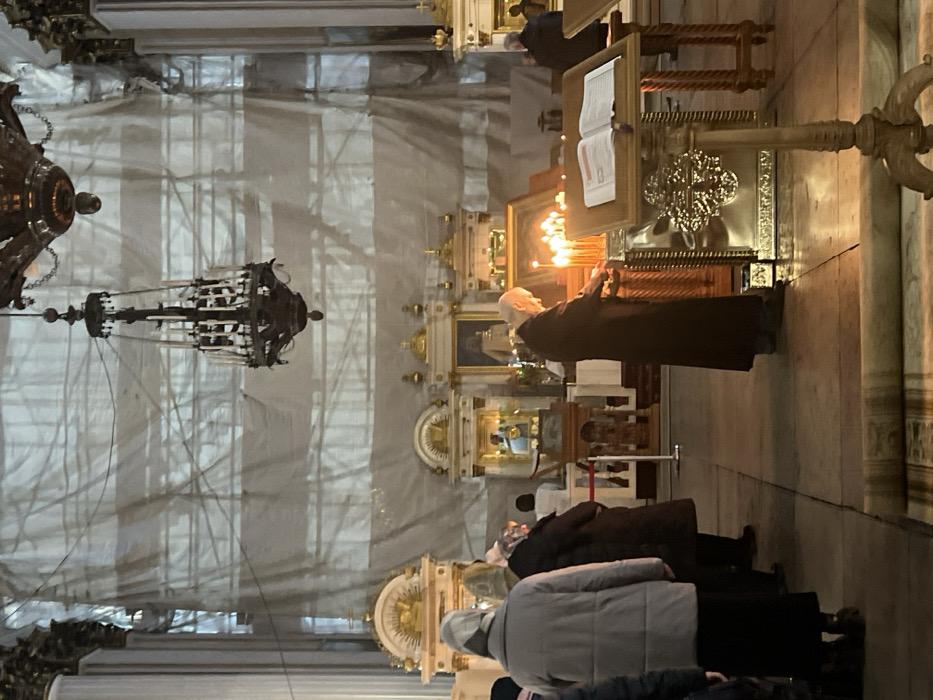 The image depicts the interior of a large Orthodox cathedral, identified as Saint Isaac's Cathedral in Saint Petersburg, Russia, based on its distinctive architectural features, including massive fluted columns, ornate gilded altars, and grand scale.
In the mid-ground, an elderly individual, dressed in a long dark robe, stands at an ornate golden candle stand and is in the act of lighting a wax candle from a cluster of already lit candles. Numerous flickering flames illuminate the immediate area around the stand. To the right of the candle stand, a wooden lectern with an open book is positioned.
In the left mid-ground, a group of approximately three to four individuals, primarily women wearing headscarves and winter coats, stand facing away from the camera, observing the scene or engaging in prayer. One woman in a light grey coat is prominent in this group.
The background reveals multiple gilded religious structures, likely parts of an iconostasis or side altars, adorned with icons and decorative elements. The upper sections of the cathedral walls and ceiling are covered with light-colored tarpaulins or sheeting, indicating ongoing renovation or preservation work, with scaffolding lines faintly visible beneath. Two large, dark-colored chandeliers hang from the high ceiling. The floor is composed of light-colored stone tiles, with a rolled-up rug visible in the lower foreground. The overall lighting is dim, accented by the glow of the candles.