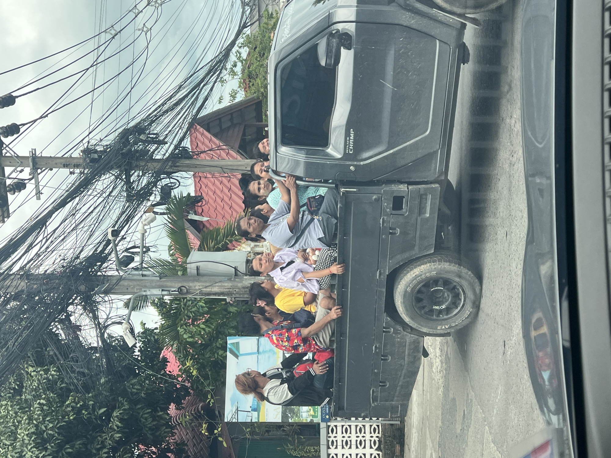 A dark-colored pickup truck, identified by the "HILUX CHAMP" text on its door, is loaded with at least eight people riding in the bed of the truck. The individuals are of various ages and genders, sitting closely together on the truck bed. In the foreground, a large utility pole is densely covered with a complex network of electrical and telecommunication wires. The background shows residential buildings with red tiled roofs and lush green trees. The scene appears to be on a paved road in a town or city, consistent with a location like Ban Bang Rak, Thailand, given the architecture and vegetation. The sky is overcast.