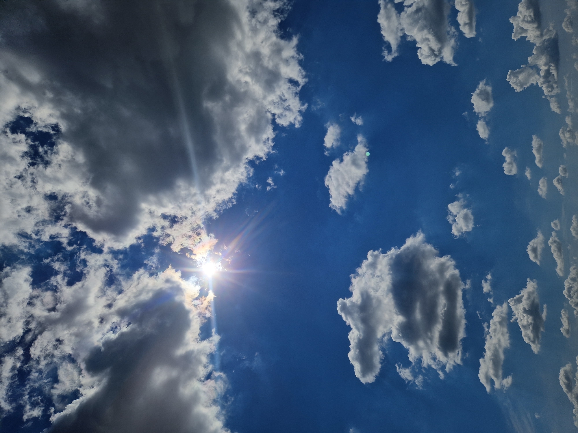 The image captures a dynamic sky filled with a mix of bright white and dark gray clouds against a vivid blue backdrop. The sun is prominently featured, peeking through a dense cluster of clouds on the left side of the frame, casting brilliant rays and creating a sunburst effect with rainbow-like halos. The clouds are varied in size and texture, from wispy cirrus formations to large, puffy cumulus clouds, some of which appear quite dense and shadowed, suggesting a change in weather.
The orientation of the image is such that the sky appears to be seen from a low angle, perhaps looking upwards from the ground. The lighting suggests it is daytime, with the bright sun indicating it could be midday or late afternoon. There are no people or man-made objects visible, focusing solely on the natural phenomenon of the sky. The overall impression is one of natural beauty and power, with the interplay of light and shadow on the clouds creating a sense of depth and drama. The presence of both bright and dark clouds hints at a day with changing weather conditions. The provided location context of Ustyvytsia, Ukraine, places this scene within a specific geographical setting, though no overt visual cues in the image directly confirm this location beyond the general appearance of a temperate climate sky.