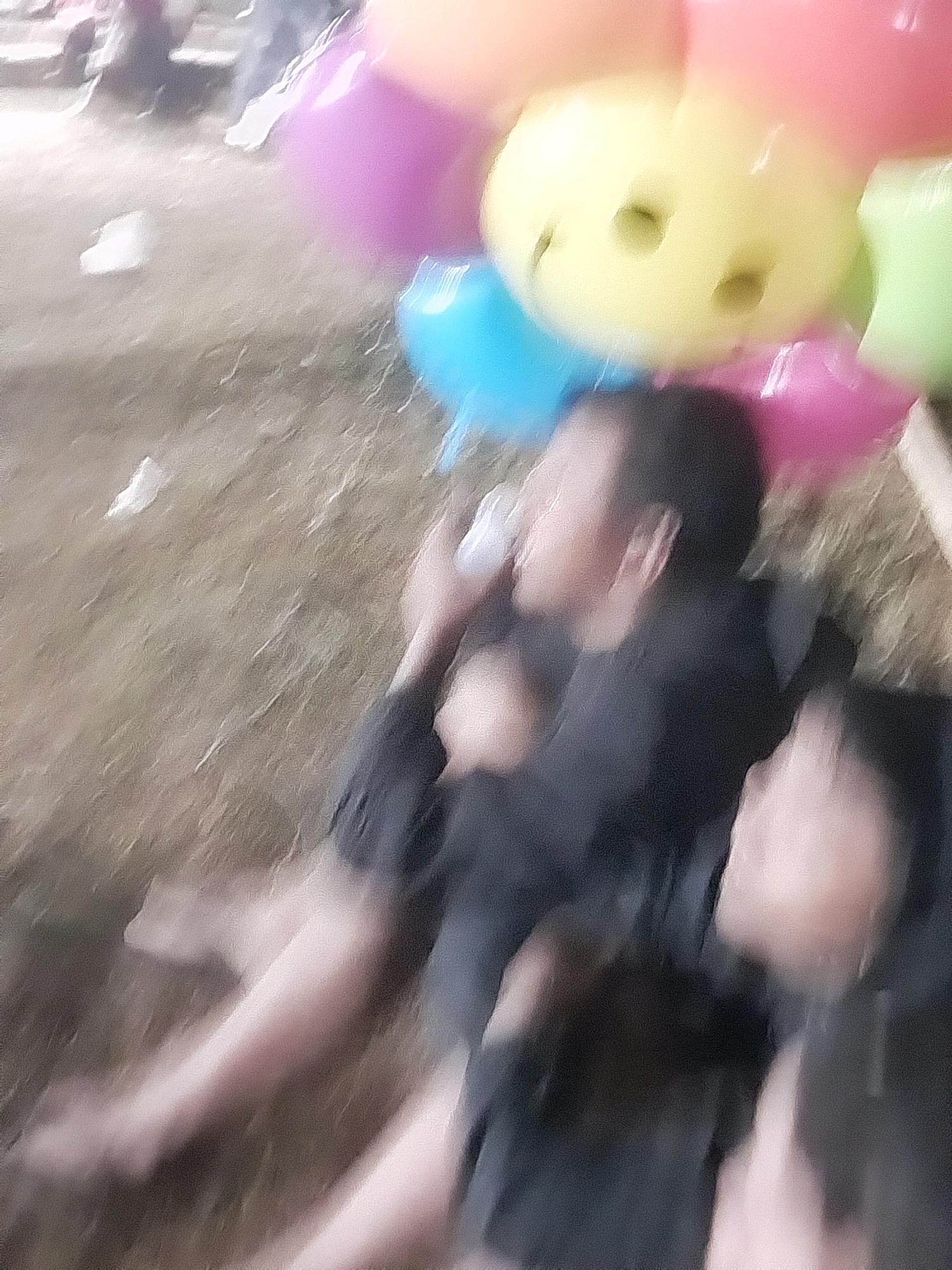 In a blurred outdoor scene, possibly in the city of Nglegok, Indonesia, several people are present near a display of colorful balloons. In the foreground, two individuals are seated or lying on the ground. The person on the left is wearing a dark shirt and has their arm extended. Their face is turned away from the camera and appears to be in profile, with their ear visible. The person to their right is also wearing dark clothing, and their head is tilted downwards, with their face partially obscured.
Behind these individuals, a collection of brightly colored balloons is visible, including pink, purple, yellow, and blue. The yellow balloon in the center has two dark indentations that resemble eyes, suggesting it is shaped like a smiley face. The balloons appear to be floating and are arranged in a cluster. The ground is covered with what looks like dry grass or soil. The overall image is out of focus, giving it a dreamy or abstract quality. The lighting is uneven, with some areas appearing brighter than others, contributing to the blurred effect.