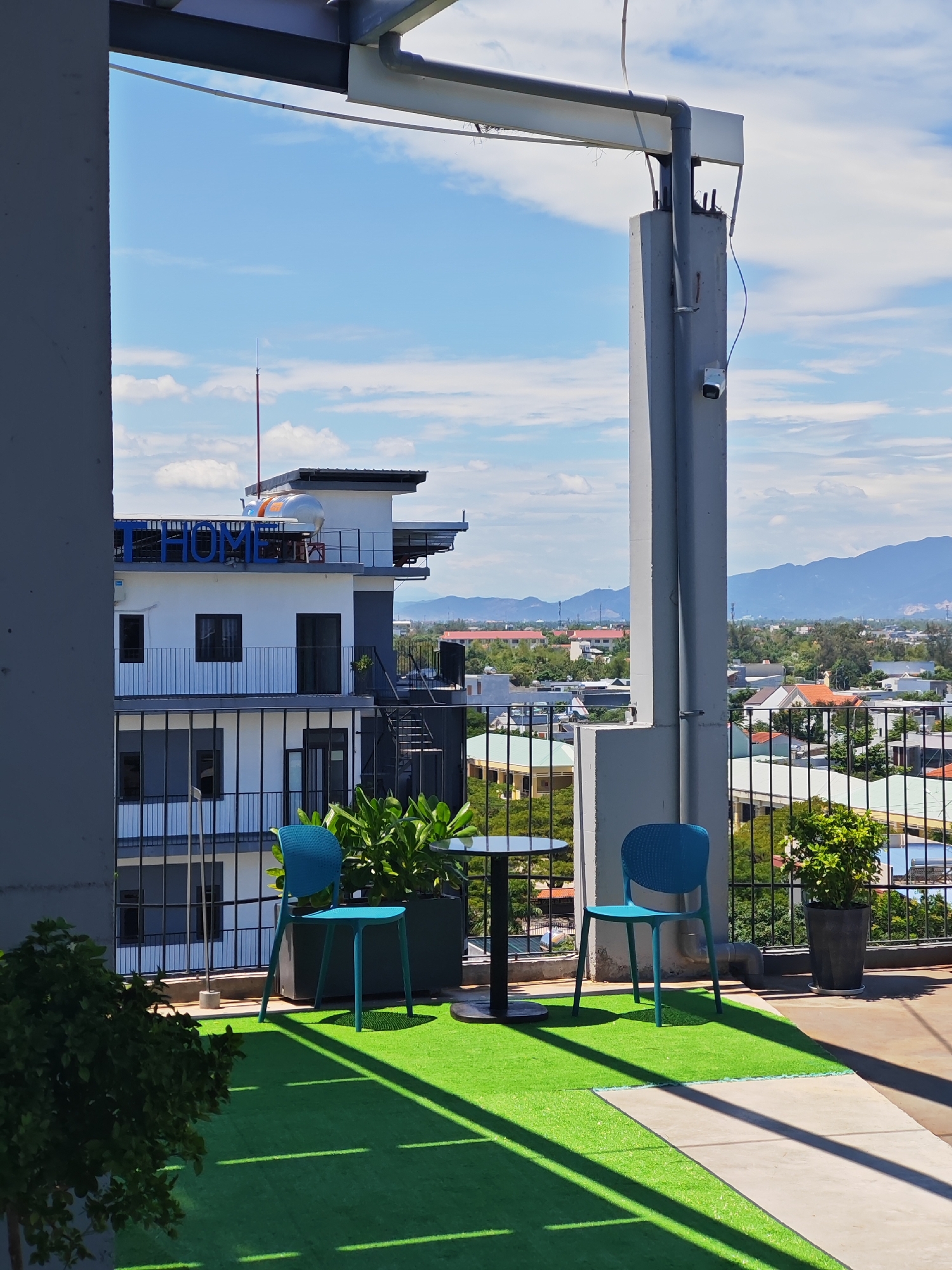 This is a slightly elevated outdoor shot of a balcony in Da Nang, Vietnam, with a view of the city and surrounding hills. The foreground features a bright green artificial turf floor with distinct shadows cast from unseen objects. Two blue chairs and a small black table are arranged on the turf. In the mid-ground, a modern white building with dark accents is visible, featuring balconies with black railings and what appears to be signage on its rooftop. Beyond the building, a cityscape unfolds with numerous residential structures and lush greenery. In the distance, a range of mountains forms the horizon under a partly cloudy blue sky. A security camera is mounted on a concrete pillar in the right mid-ground.