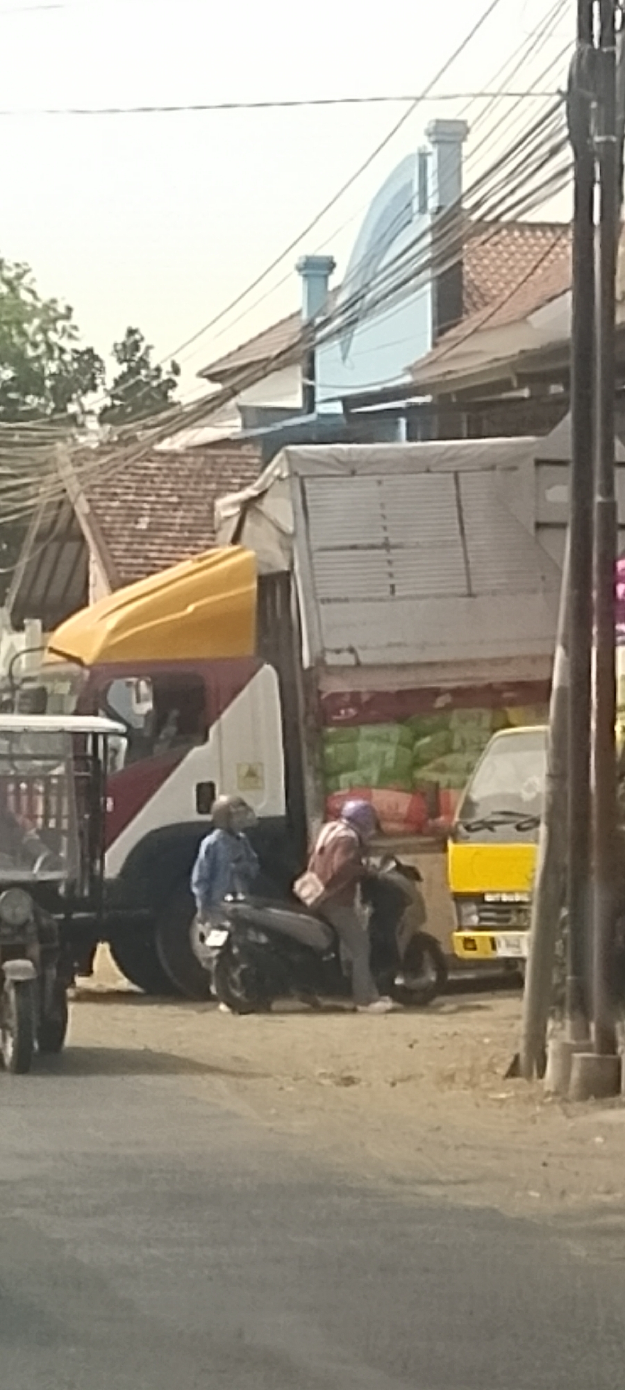 In Panjang, Indonesia, two individuals on a motorbike pause beside a large truck. The truck, a white and red cargo vehicle, has its rear tarp pulled up, revealing stacked goods, possibly sacks of produce. The truck's yellow cab is visible. To the left of the truck, a smaller, white vehicle with a canopy, resembling a three-wheeled transport, is parked. The person in the foreground on the motorbike wears a blue jacket and a helmet. The passenger behind wears a brown jacket and a purple helmet. They appear to be stopped on a dusty, unpaved area beside a road. In the background, buildings with red-tiled roofs and overhead power lines suggest an urban or semi-urban setting. The sky is bright, indicating daylight.