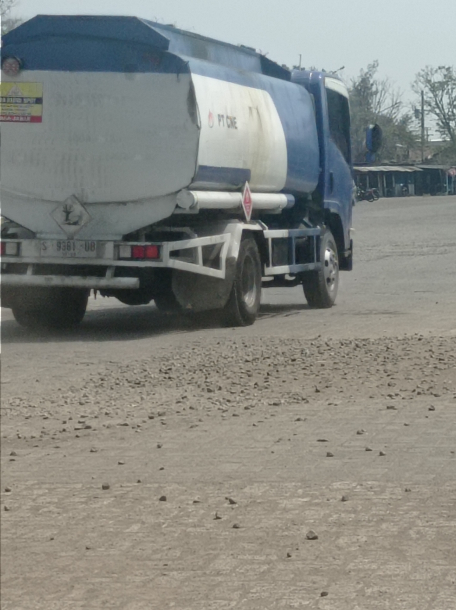 A blue and white tanker truck is parked on a dusty, unpaved surface in Tegal, Indonesia. The truck has "PT CNE" written on its side. A hazard symbol, which appears to be a fire hazard, is visible on the rear of the truck. The truck is facing away from the viewer and appears to be stationary. In the background, there are several simple buildings with corrugated metal roofs, suggesting a commercial or industrial area. Bare trees with thin branches are visible against a hazy sky. The ground is covered in loose gravel and dirt.
