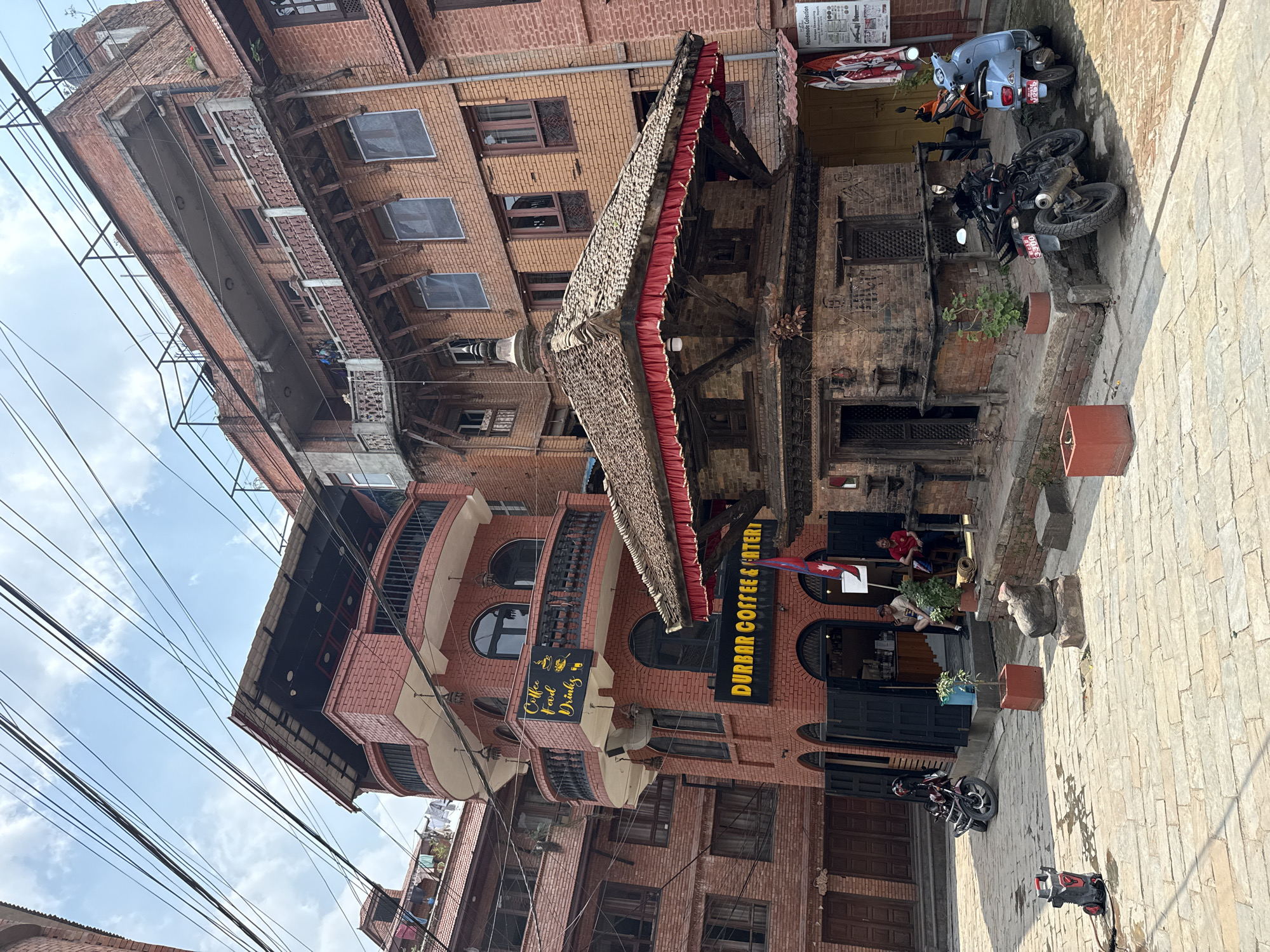 This image shows the Darbar Coffee & Eatery located in Bhaktapur, Nepal. The eatery is housed in a red brick building with arched windows and a thatched awning. The street in front of the building is paved with bricks. There are several motorcycles parked near the cafe. People are sitting at tables outside the eatery. The surrounding buildings are constructed of brick. Overhead are numerous wires. The sky is partially cloudy.