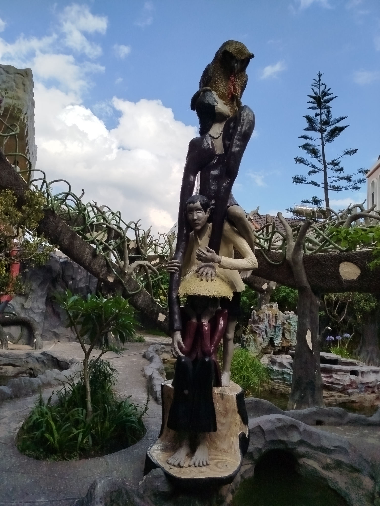 The image depicts a multi-figure sculpture in what appears to be a garden or park setting. The sculpture is composed of three figures stacked on top of each other, seemingly forming a human tower. The base figure is sitting, the middle figure is holding the second one on their shoulder. The top figure has a large bird perched on the head. The sculpture stands on a tree trunk base. The surrounding environment includes trees, foliage, and textured stone or concrete structures designed to mimic natural rock formations. In the background, there is a pine tree and buildings. Based on visual characteristics, this sculpture is situated within the grounds of Crazy House in Da Lat, Vietnam.
