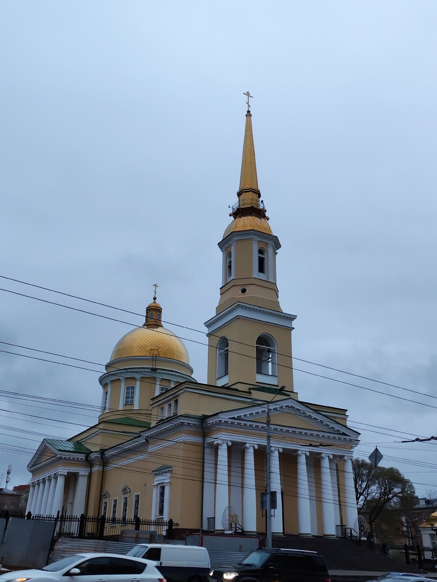 The image shows the Alexander Nevsky Cathedral in Izhevsk, Russia, set against a cloudy sky. The cathedral has a yellowish-orange facade with white columns supporting a portico. Two golden domes are visible; a larger one to the left and a taller spire to the right, both topped with Orthodox crosses. Power lines run across the foreground of the image. Several cars and a van are partially visible at the bottom of the frame, suggesting the cathedral is located along a street. The street appears to have a staircase leading up to the building.