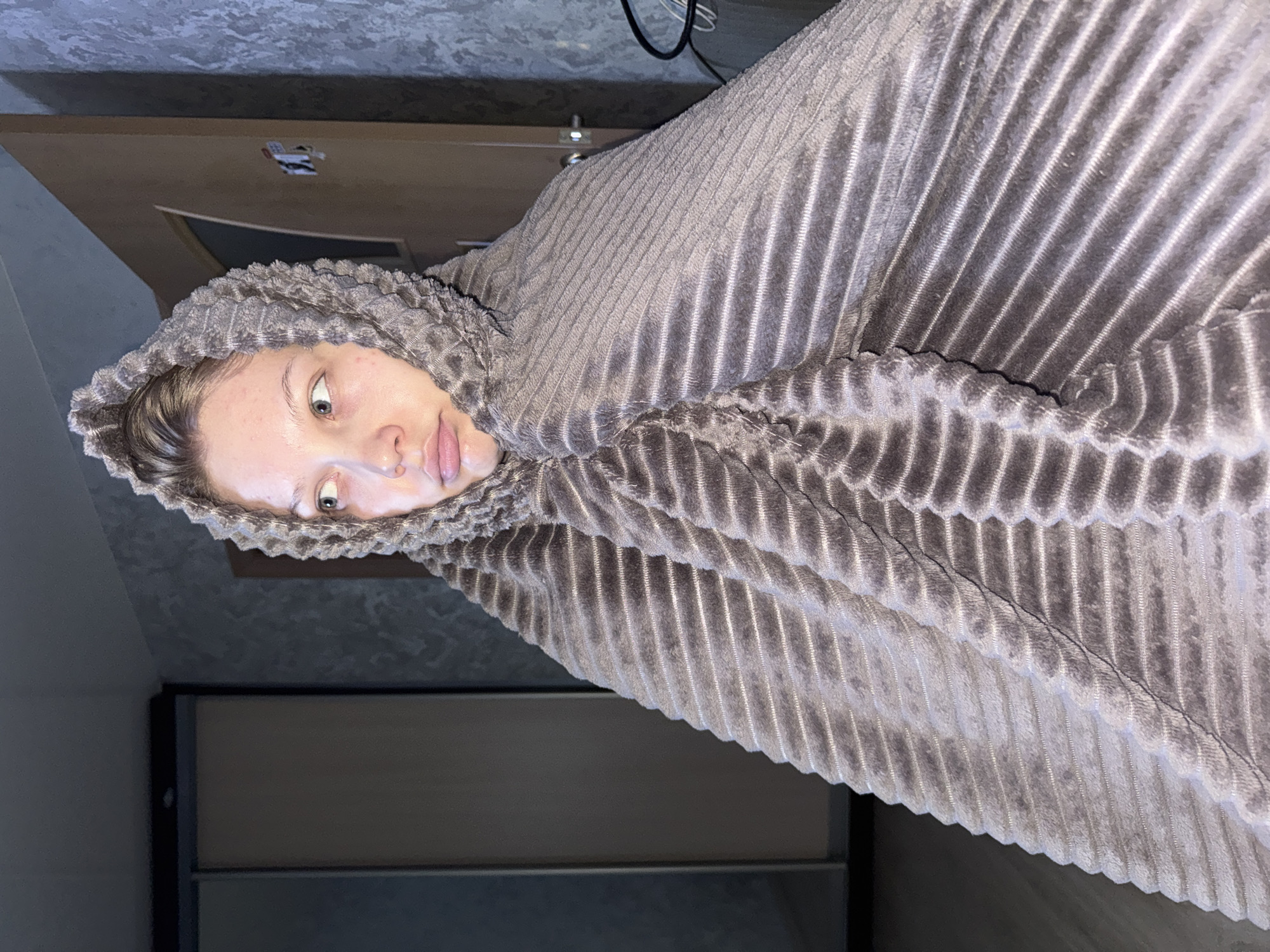 A person is wrapped in a gray, ribbed, hooded blanket. The person has fair skin and is looking at the camera. The location appears to be indoors. Elements in the background include a door, a piece of furniture, and a ceiling. The image does not provide information to confirm the location is Chaikovsky, Russia.