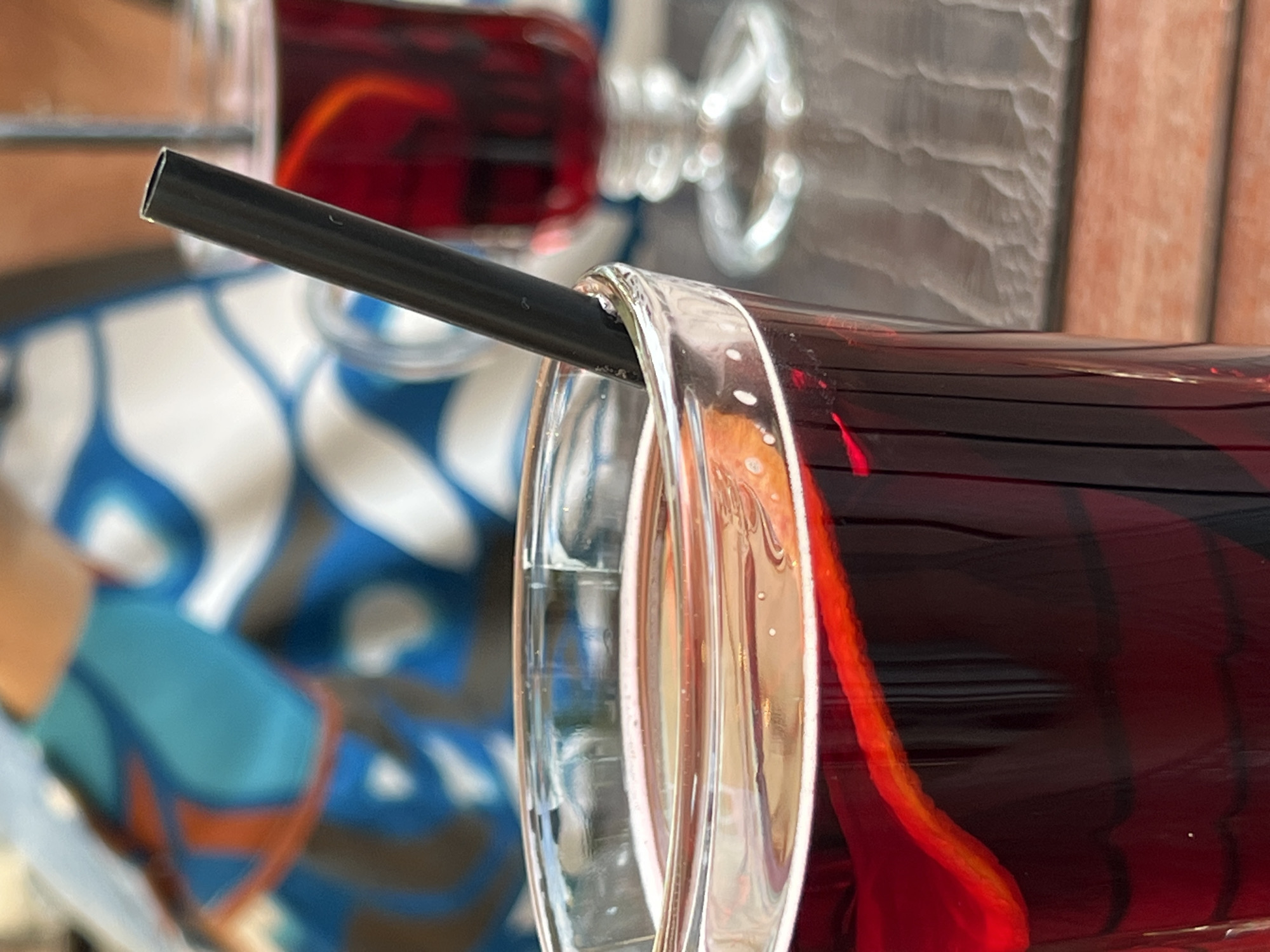 A close-up shot focuses on a clear glass filled with a dark red beverage, likely wine or a similar drink, with a slice of orange floating near the rim. A black straw protrudes from the glass. In the blurred background, another glass of the same red beverage is visible on a table, along with a person wearing a blue and white patterned top. The scene appears to be an outdoor setting, possibly a cafe or restaurant, with a wooden table in the foreground and a hint of a patterned surface in the background. The lighting suggests it could be daytime. The photo's composition suggests it was taken in Palanga, Lithuania, a popular seaside resort town known for its cafes and relaxed atmosphere.