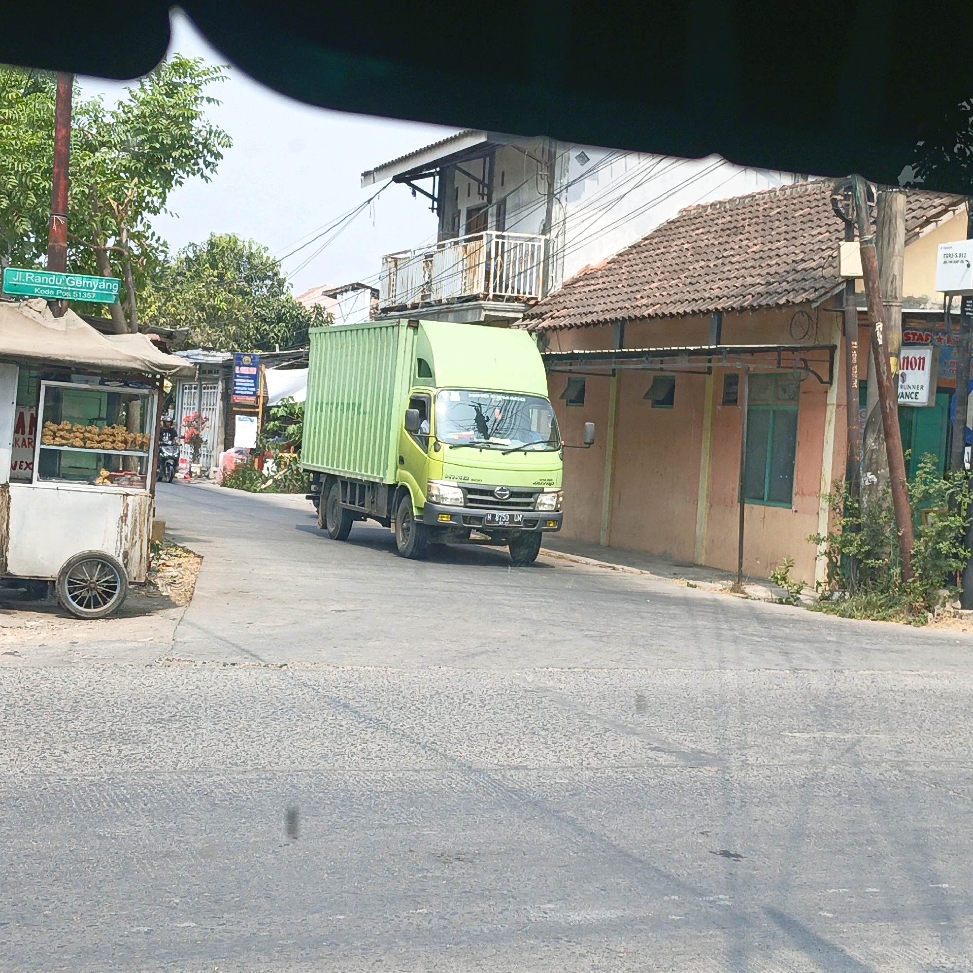 A light green truck with a large box trailer is driving on a paved road in Pegandon, Indonesia. The truck has a license plate that reads "H 8750 LM". To the left of the truck, a street food cart is parked on the side of the road. The cart is white with a canvas awning and displays fried foods in a glass case. A person is visible riding a motorcycle in the background, heading away from the viewer. Buildings with tile roofs line the street. A street sign indicates "Jl. Randu Gembyang" and "Kode Pos 51357". The environment appears to be a commercial or residential area with moderate vegetation.