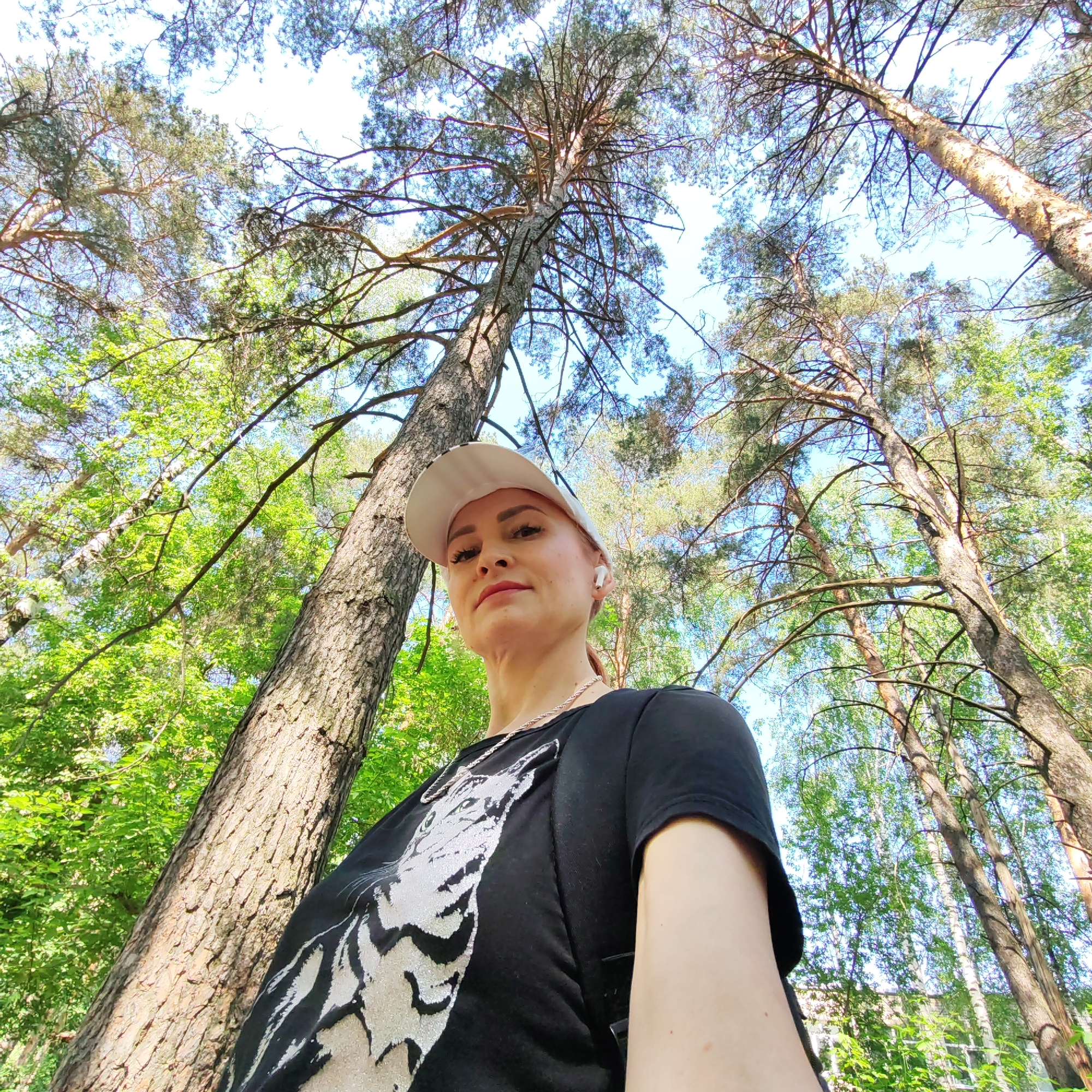 A low-angle shot shows a woman wearing a white cap, black t-shirt with a light-colored animal design, and an earbud in her right ear. She stands in a forest with tall trees. The tree to her left is in focus, while the background displays a mix of tall pine trees and green-leaved trees against a partly cloudy sky. The location is Chaikovsky, Russia.