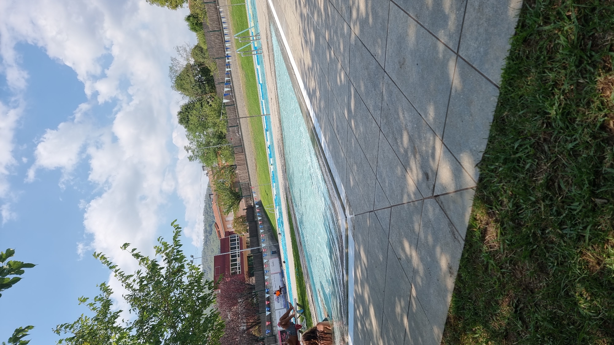 The image shows an outdoor swimming pool facility in Sant Joan Les Fonts, Spain. The foreground features a paved area with large square tiles and a strip of green grass. Beyond this, a large, bright blue swimming pool with clear water occupies the middle ground. Several people are visible in and around the pool. One person is partially submerged in the water, and another is lying on a lounger. In the background, there is a grassy area with a fence, and beyond that, buildings and trees. The sky is blue with scattered white clouds, and sunlight casts shadows on the paved area. The overall impression is a sunny day at a recreational facility.