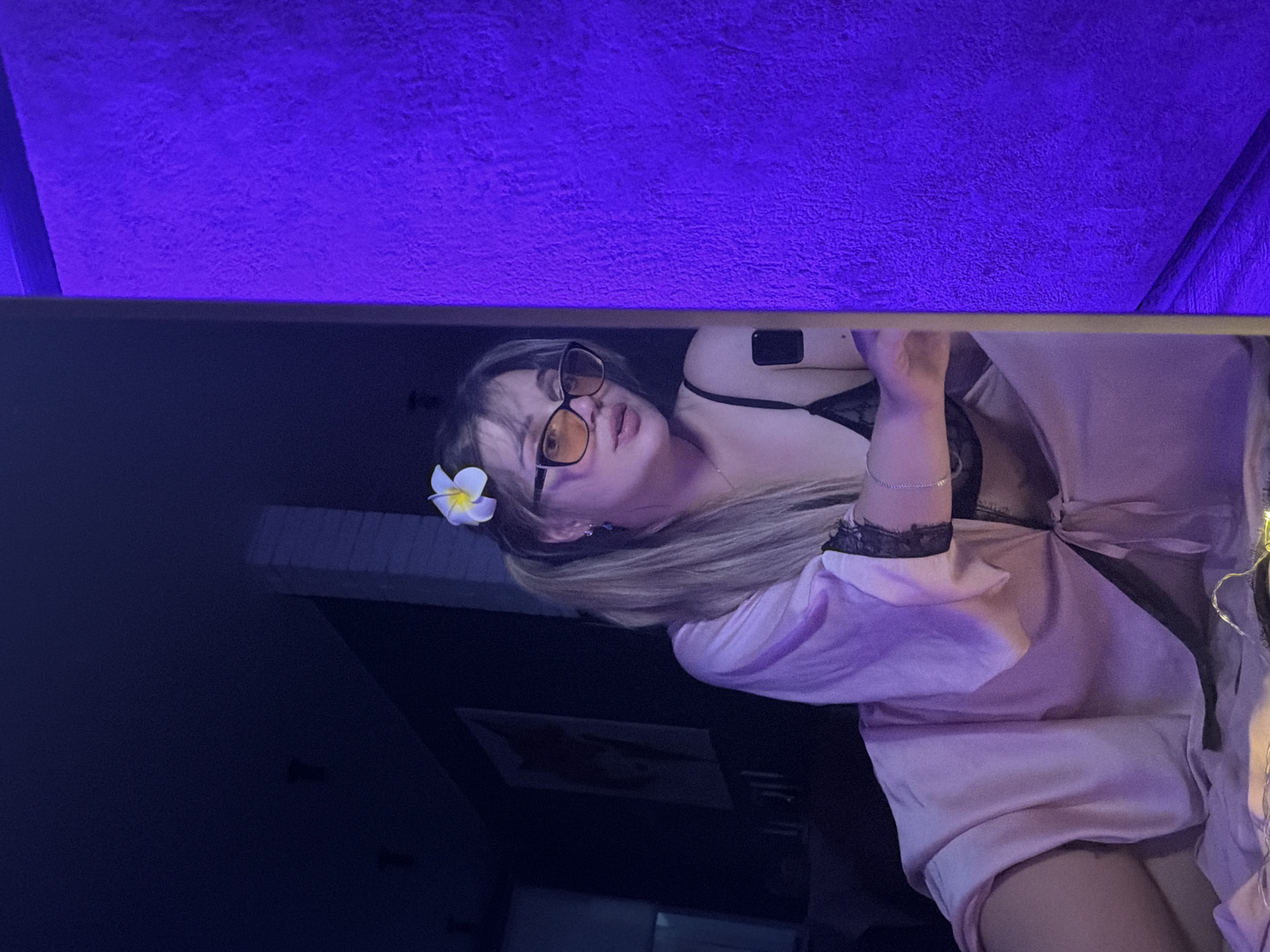 A woman is lying on a bed or other surface. She is wearing a light pink robe over what appears to be a black lace top or bra. She has a white flower in her hair and is wearing glasses with yellow-tinted lenses. The background appears to be a dark room, illuminated by blue lighting. A textured blue ceiling or wall is visible in the upper portion of the frame. There's a photo on the wall behind her. I do not have enough information to know the location is Большая Шильна, Russia.
