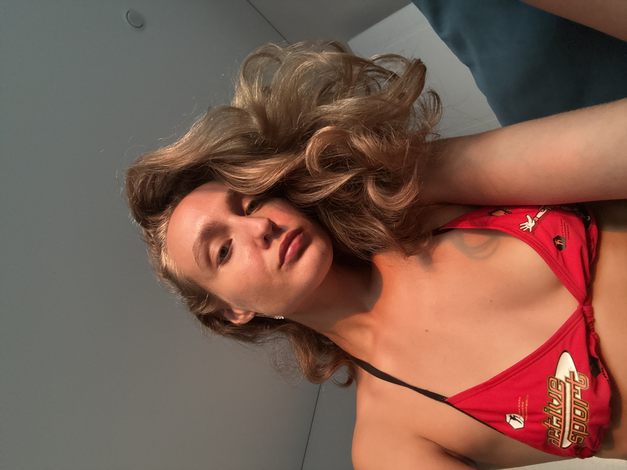A low-angle, close-up, selfie-style photograph shows a young woman from the chest up, looking directly at the camera. She has long, wavy, light brown hair that is styled to the side. Her skin is lightly tanned, and she is wearing a red bikini top with a black string tie. The bikini top has white lettering that appears to say "active sport" and other graphics. The lighting suggests the photograph was taken indoors near a window, with sunlight illuminating her face and upper body from the side. The background consists of a plain, light gray ceiling and a corner of a teal-colored cushion or piece of furniture. The image does not contain any specific details indicating a location within the city of Подшивалово, Russia.