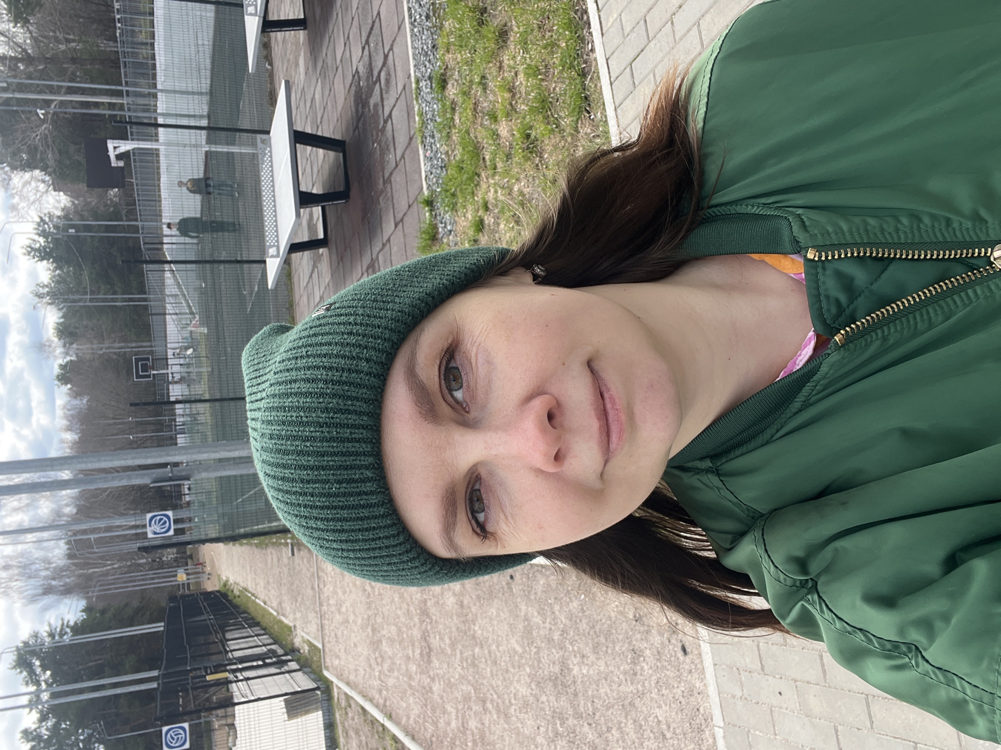 The image is a vertical selfie featuring a young woman in an outdoor setting in Chaikovsky, Russia. She is positioned in the foreground, looking directly at the camera with a subtle, pleasant smile. She has long, dark brown hair and green eyes, and is wearing a green knit beanie and a matching green bomber jacket, with a colorful (orange and pink) garment visible beneath.
The background shows an outdoor sports complex. Two white outdoor ping-pong tables with dark frames are prominent in the mid-ground. Behind these, a sports court is enclosed by a metal mesh fence. A basketball hoop with a backboard is visible on the court, where two blurry figures (possibly men) are standing. Two white circular signs with a blue volleyball logo are attached to the fence. The ground is a mix of paved areas and sparse grass, with bare trees in the distance. The lighting is bright and even, indicative of an overcast day, suggesting late winter or early spring.