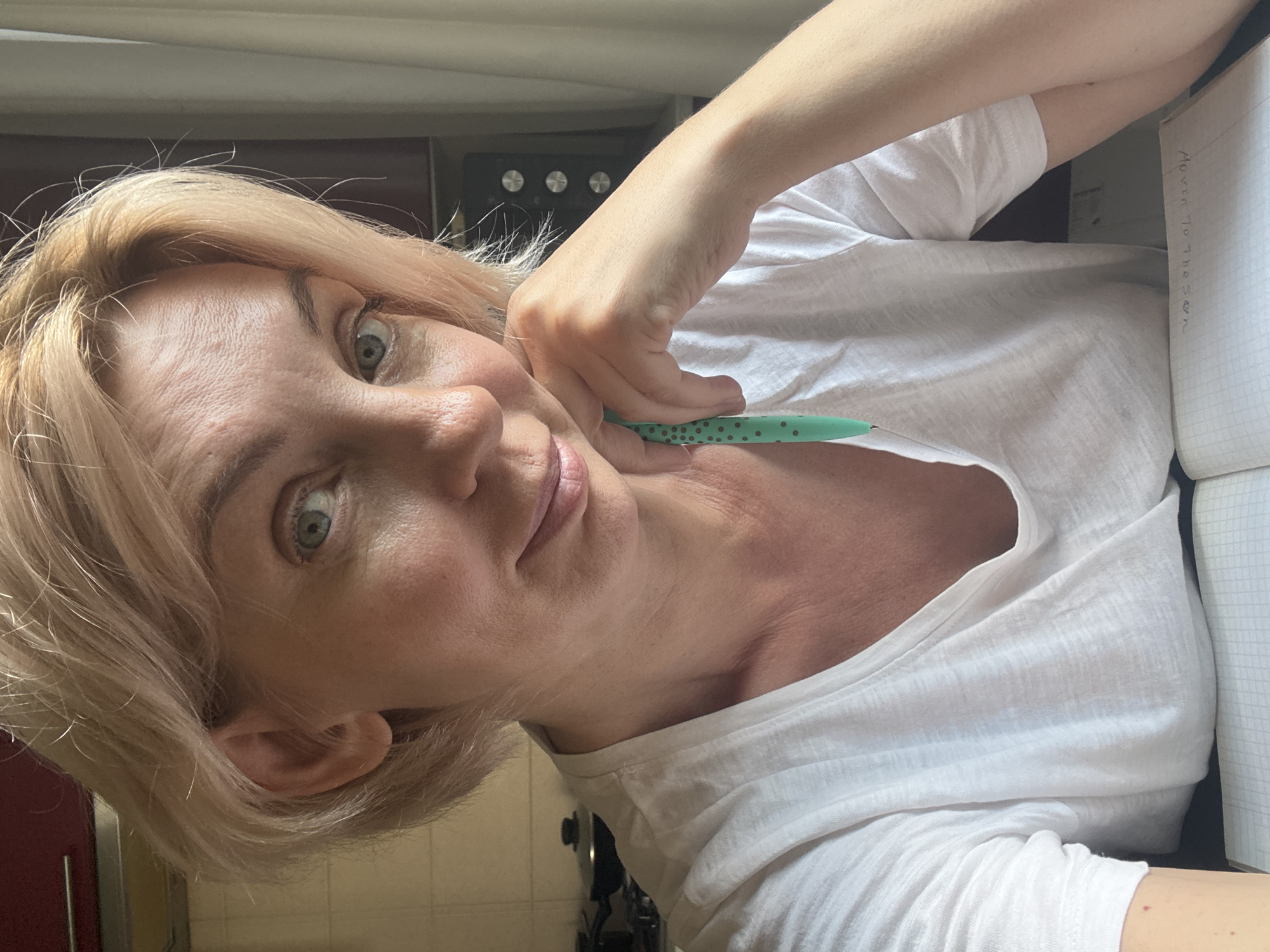 The image shows a close-up, selfie-style shot of a woman with short, blonde hair and green eyes, sitting at a table indoors. She is wearing a white t-shirt and has her right hand resting on her chin, holding a teal pen with dark green polka dots. Her left hand is out of frame. A notebook with a grid pattern is visible on the table in front of her, with some writing on it. The background includes a kitchen counter with a black stovetop, a microwave oven, and a sheer curtain. The lighting suggests it is daytime. The image does not contain any visual information that would allow for determining the location as Sochi, Russia.