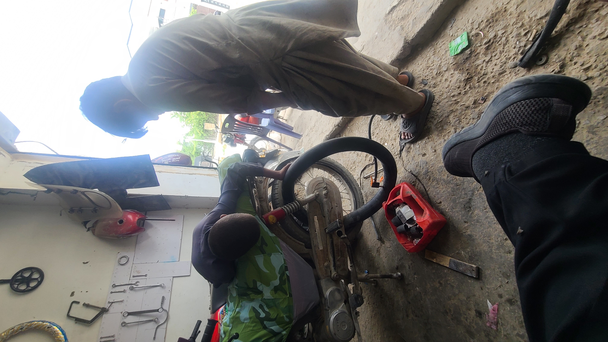 The image depicts a bustling daytime scene inside a motorcycle repair workshop, likely located in Quetta City Tehsil, Pakistan. Two men are actively engaged in working on a motorcycle, while the leg of a third individual is prominently visible in the foreground, suggesting a first-person perspective from someone observing or waiting.
One man, dressed in a light-colored traditional garment (possibly a shalwar kameez), stands bent over the motorcycle, appearing to focus on its front section or engine. The second man, wearing a dark shirt underneath a green camouflage patterned top, is crouched low beside the motorcycle's rear wheel, which seems to be detached or in the process of repair. The leg and foot of the third person in the foreground are clad in a black sneaker and dark pants.
The workshop floor is a rough, uneven surface of concrete or dirt, with scattered debris. On a white board mounted on the wall in the background, various tools such as wrenches and spanners are neatly arranged. A red plastic container, filled with what appear to be rubber scraps or pieces of inner tube, sits on the ground next to the motorcycle. Faintly visible on a motorcycle part in the upper background is the brand name "HONDA," along with what could be a model designation like "CD 70" or "CG 125," common motorcycle models in the region. The bright, natural lighting indicates it is daytime. The scene conveys an atmosphere of focused manual labor in a functional repair setting.