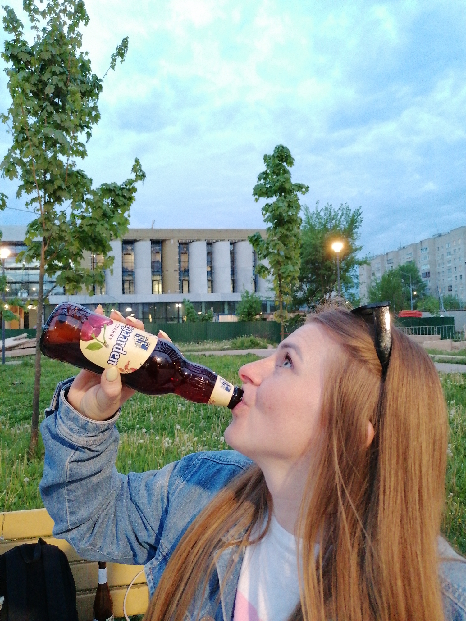 A young woman with long, straight brown hair is drinking from a brown beer bottle outdoors in a city park. She is wearing a light blue denim jacket over a white shirt and a black headband with cat ears. The beer bottle, which she is holding with her right hand, is labeled "Hoegaarden Cherry". Another beer bottle sits on the bench beside her. Behind her is a green lawn, scattered with white dandelions. Young trees are planted around the lawn. In the background is a multi-story building with white columns and rows of windows. Further back, another residential building and street lights can be seen. The sky is overcast. The location is Unknown, Unknown.