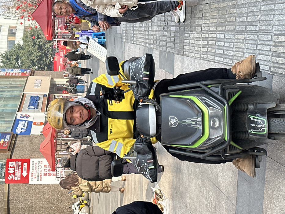 A delivery driver wearing a yellow jacket and helmet sits on a black and green motorcycle with "BLACK NIGHT" written on it. The motorcycle has a logo resembling a horse and rider. In the background, people walk along a tiled sidewalk lined with shops and vendors, including a sign advertising "over 1500 stores." The scene depicts a busy urban environment.